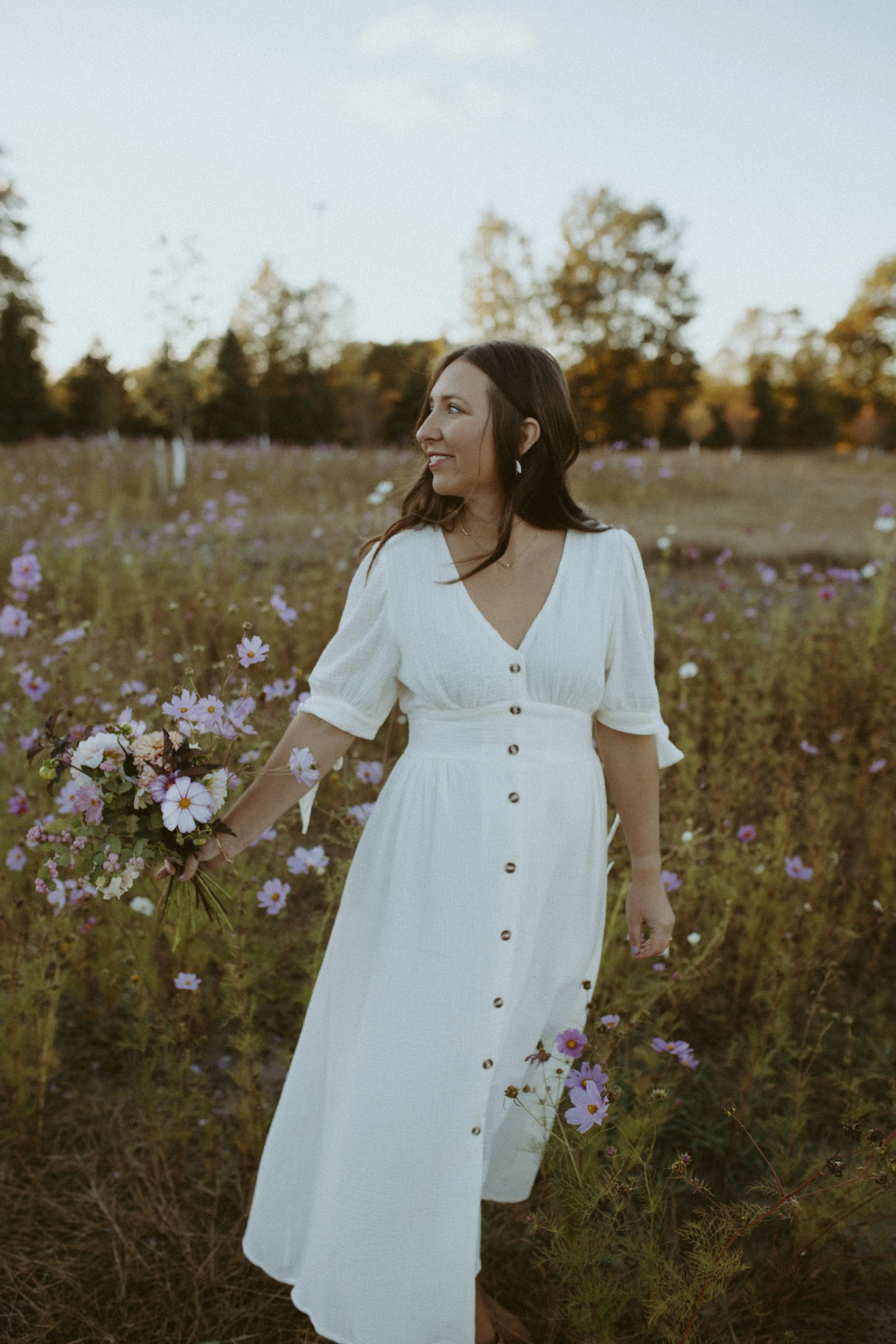 A woman in a white dress holding a bouquet of flowers standing in a field of purple flowers with trees in the background during sunset.