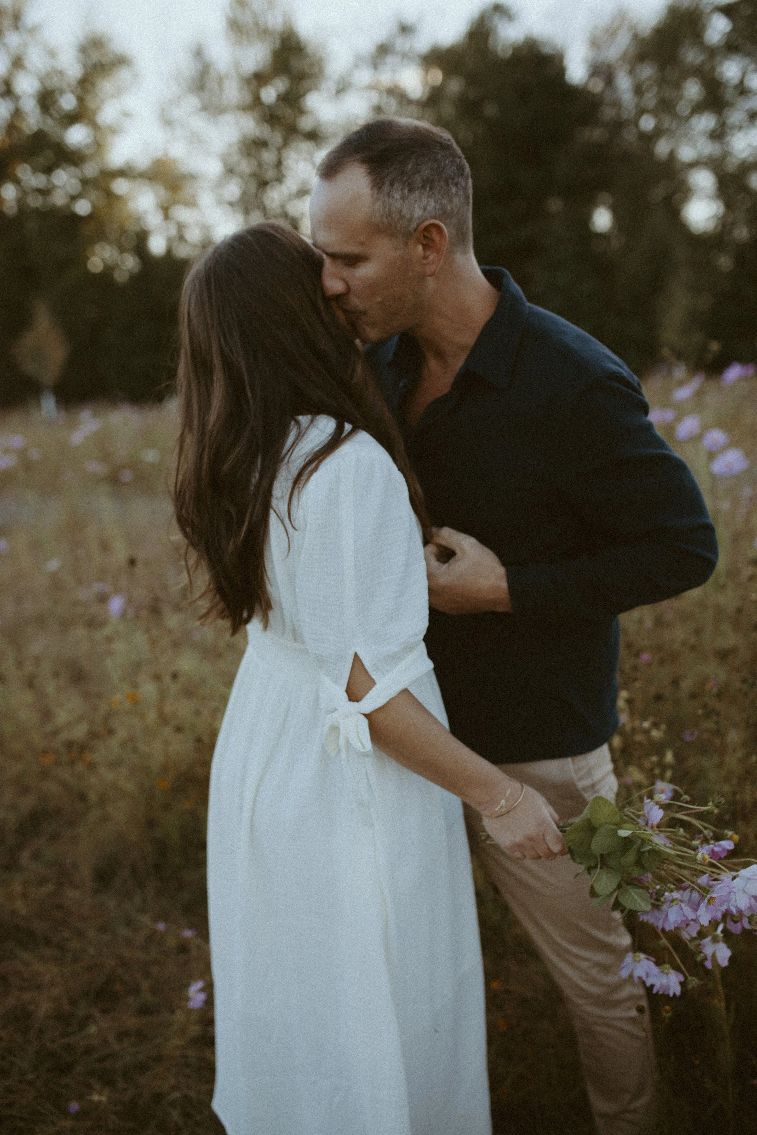 A man and woman sharing a kiss outdoors during sunset, the woman holding flowers.