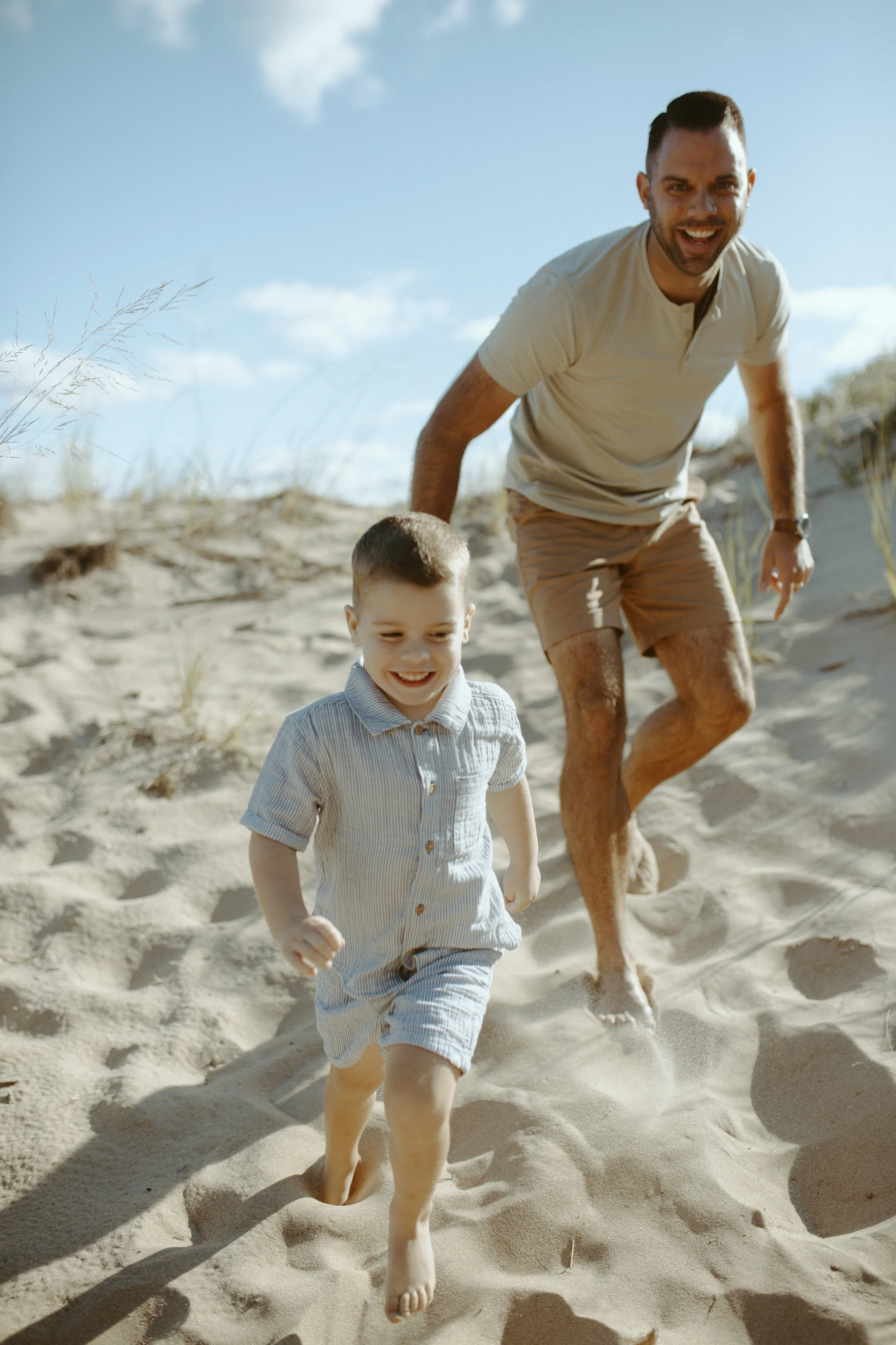 A man and a young boy running on a sandy beach under a blue sky with sparse clouds, enjoying a sunny day.