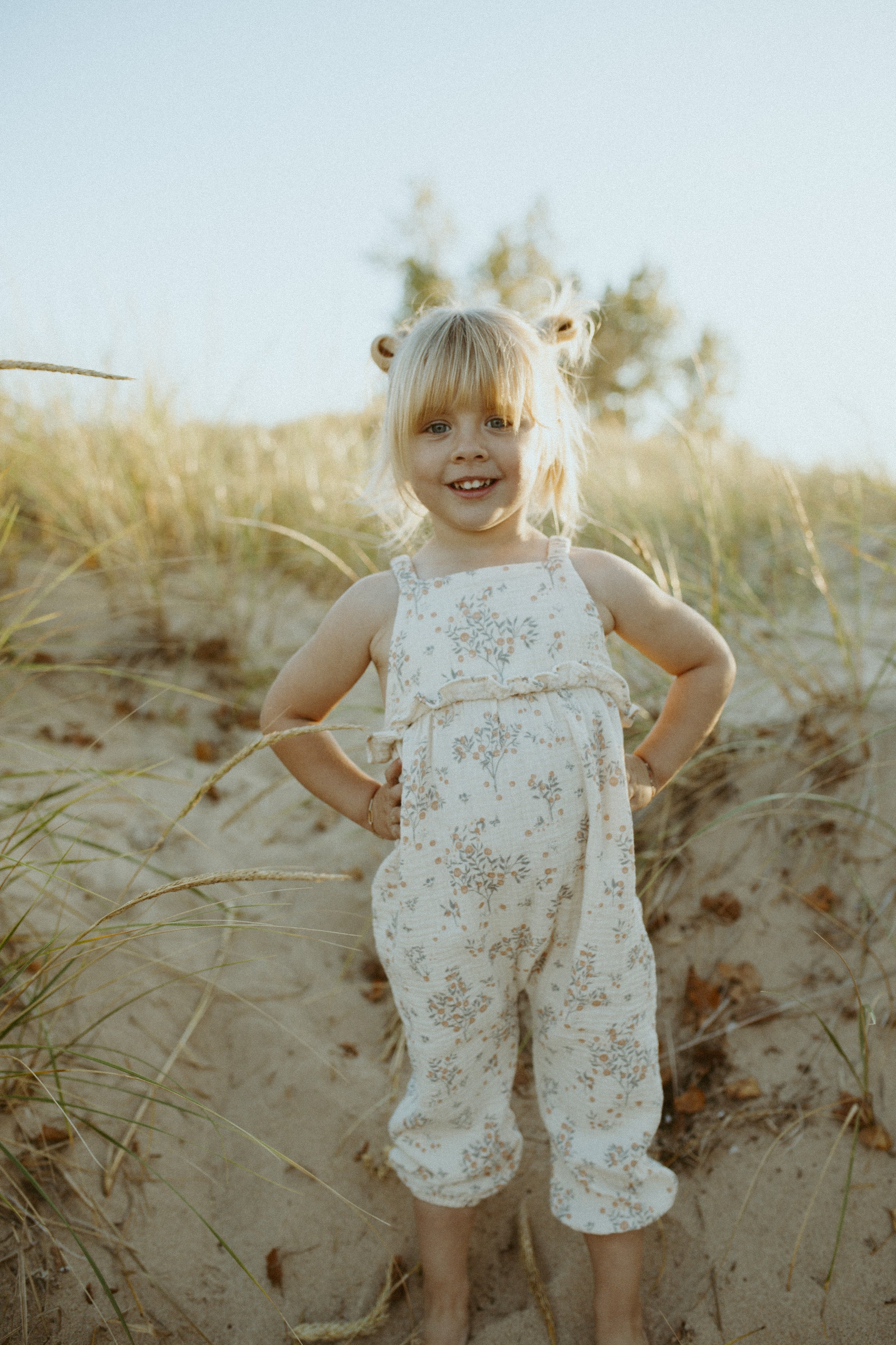 A young girl with blonde hair wearing a light-colored floral jumpsuit standing on sand with dune grass and bushes in the background, smiling and looking at the camera.
