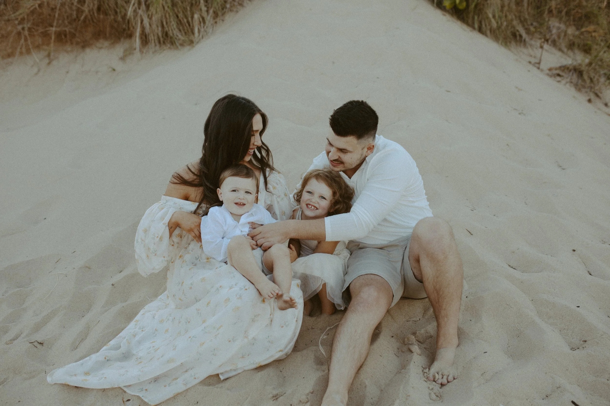 A family of four sitting on the sand at the beach, smiling and enjoying each other's company.