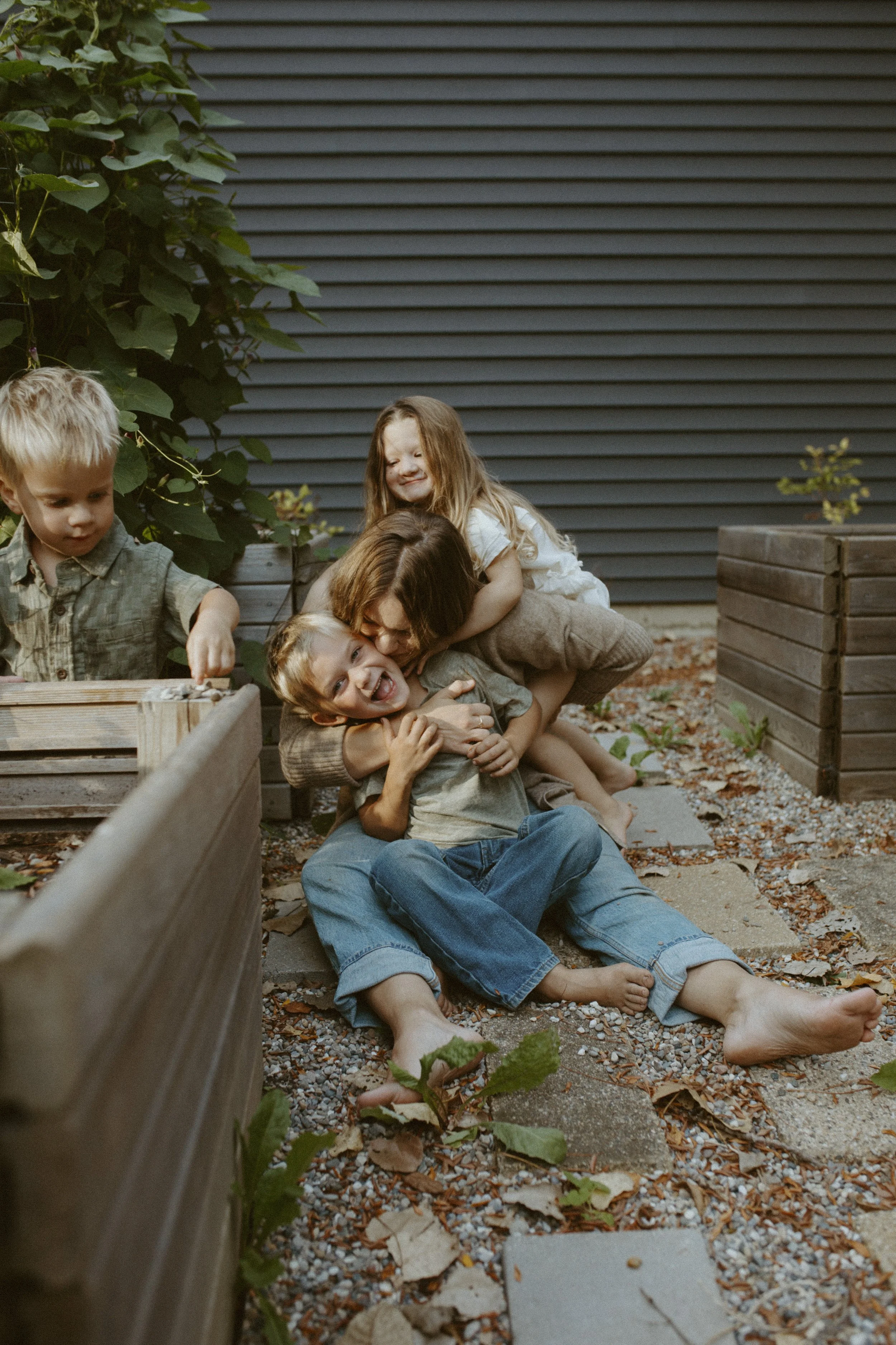 A woman and two children playing on a gravel path outside near a dark gray wall and wooden planter boxes, with some green plants and fallen leaves.
