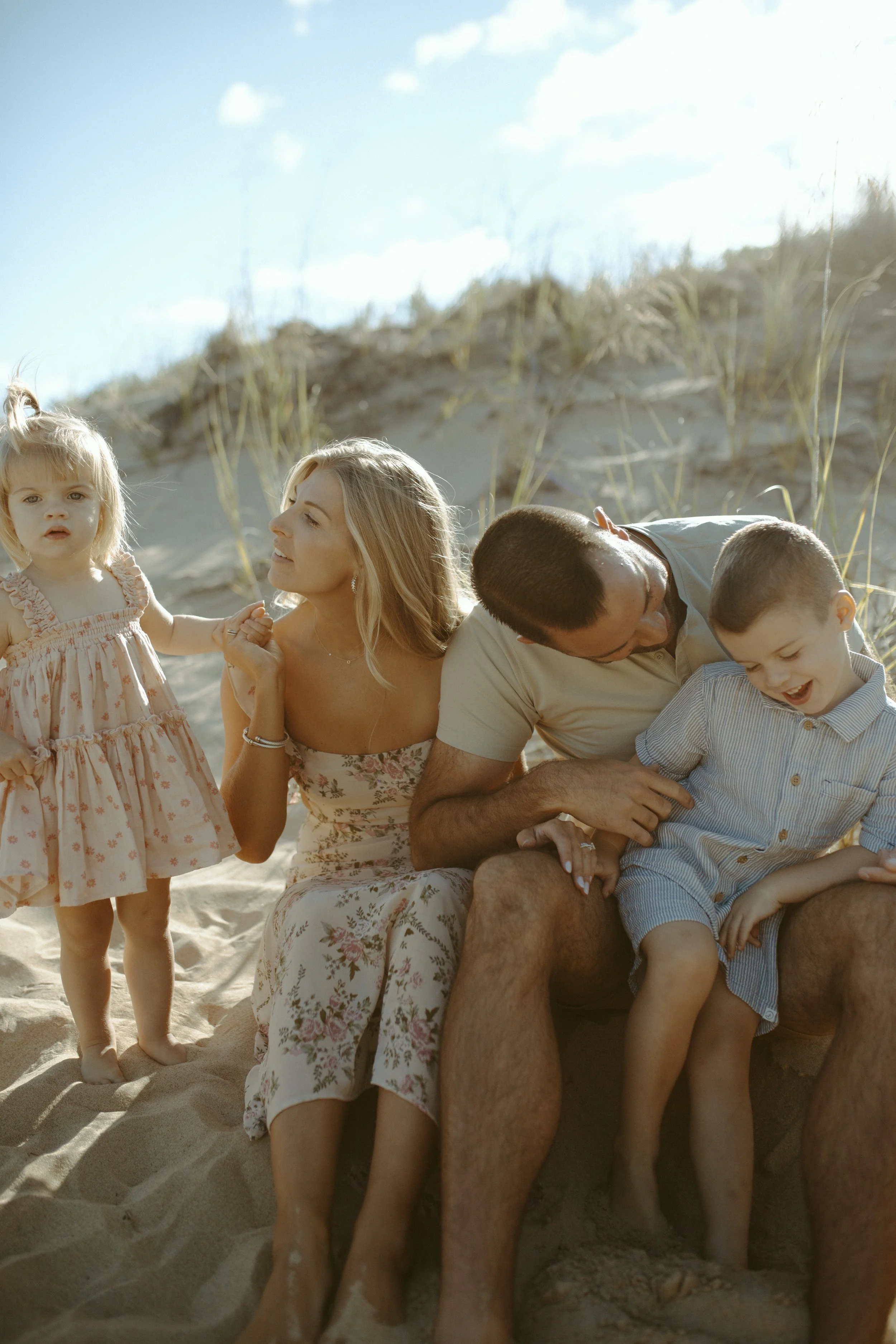 A family enjoying a day at the beach, sitting on the sand with dunes and grass in the background, smiling and playing together.