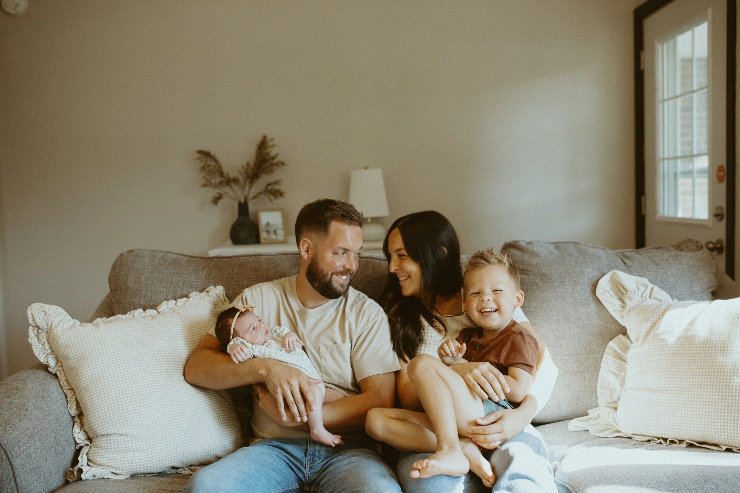 A family of five on a beige sofa in their living room, smiling and enjoying time together. The father is holding a newborn, the mother and a young boy are sitting close, and another child is next to the boy. The room has a neutral wall, a lamp, decor