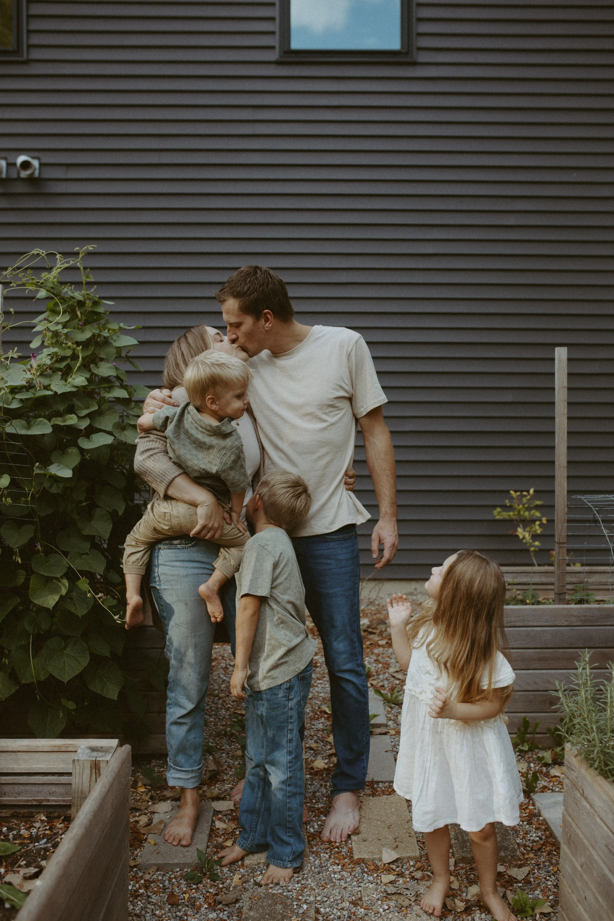 A man and woman kissing while holding children in their arms in a backyard garden. Several children stand around them, some looking up at the adults, with a house wall in the background, plants on the side, and the ground covered in leaves and gravel