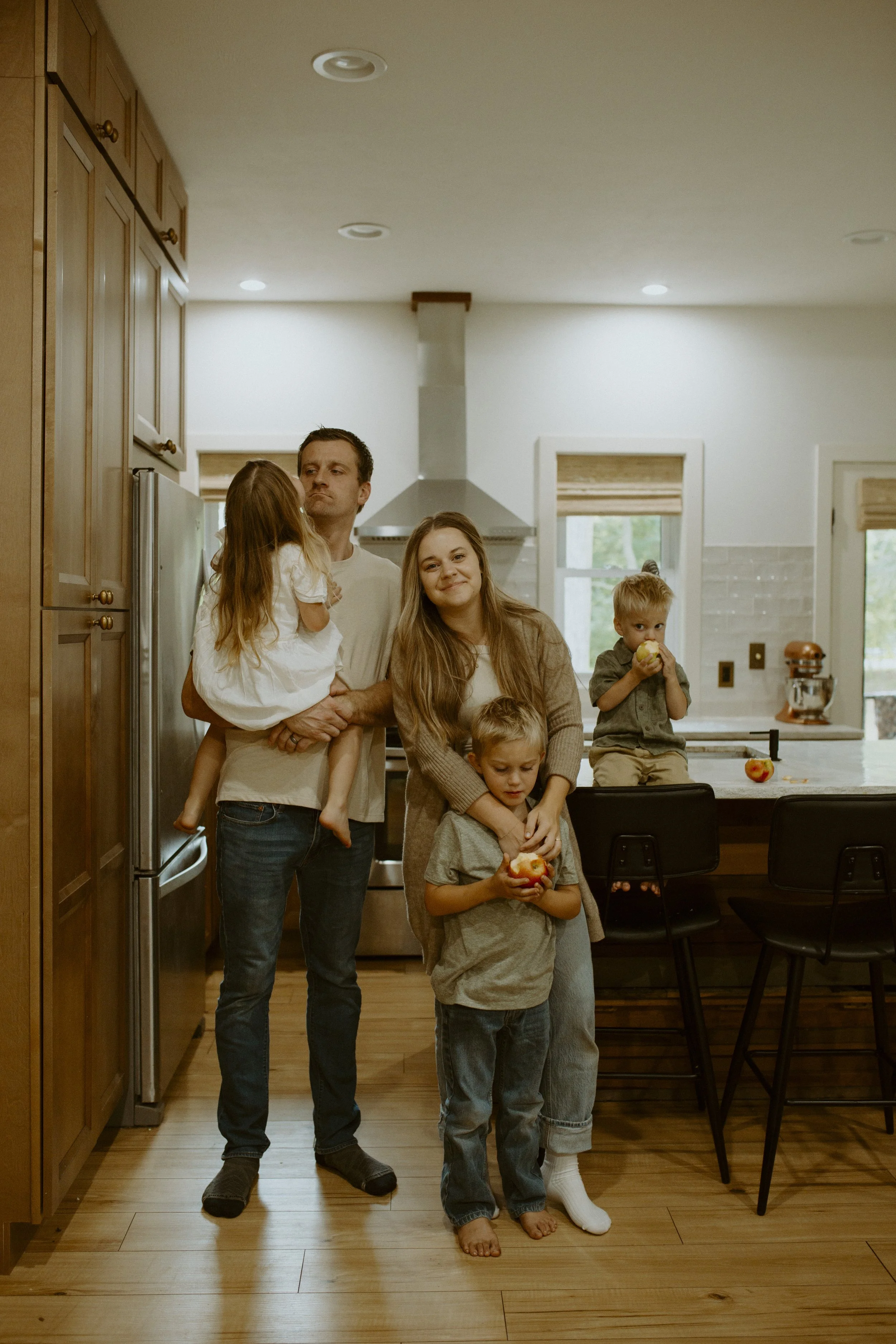 A family in a kitchen with wooden cabinets and a dining table, with two children holding apples, and a man and woman standing nearby.