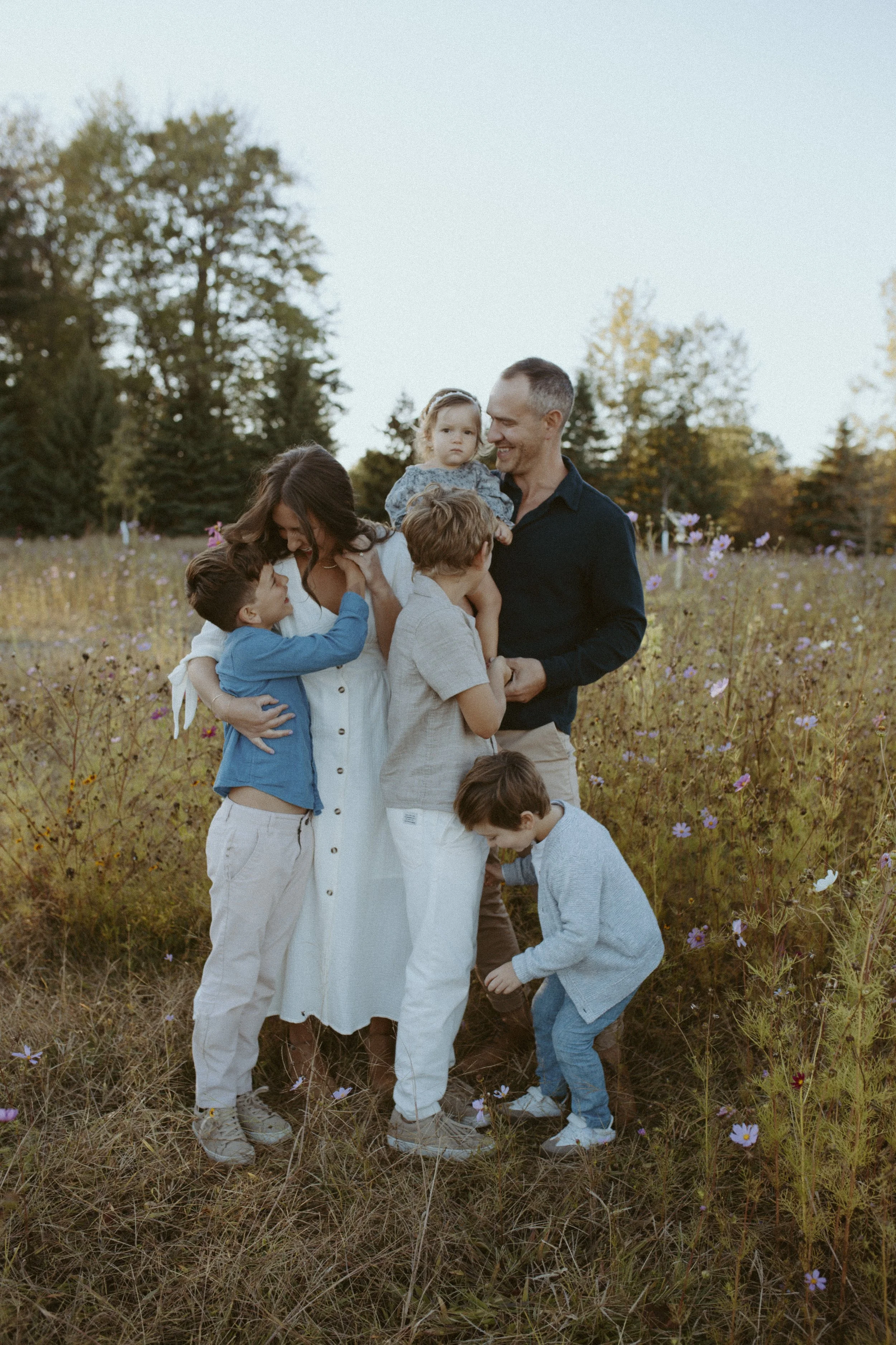 A family of six, including two adults and four children, standing in a field of flowers and tall grass with trees in the background during late afternoon or early evening.
