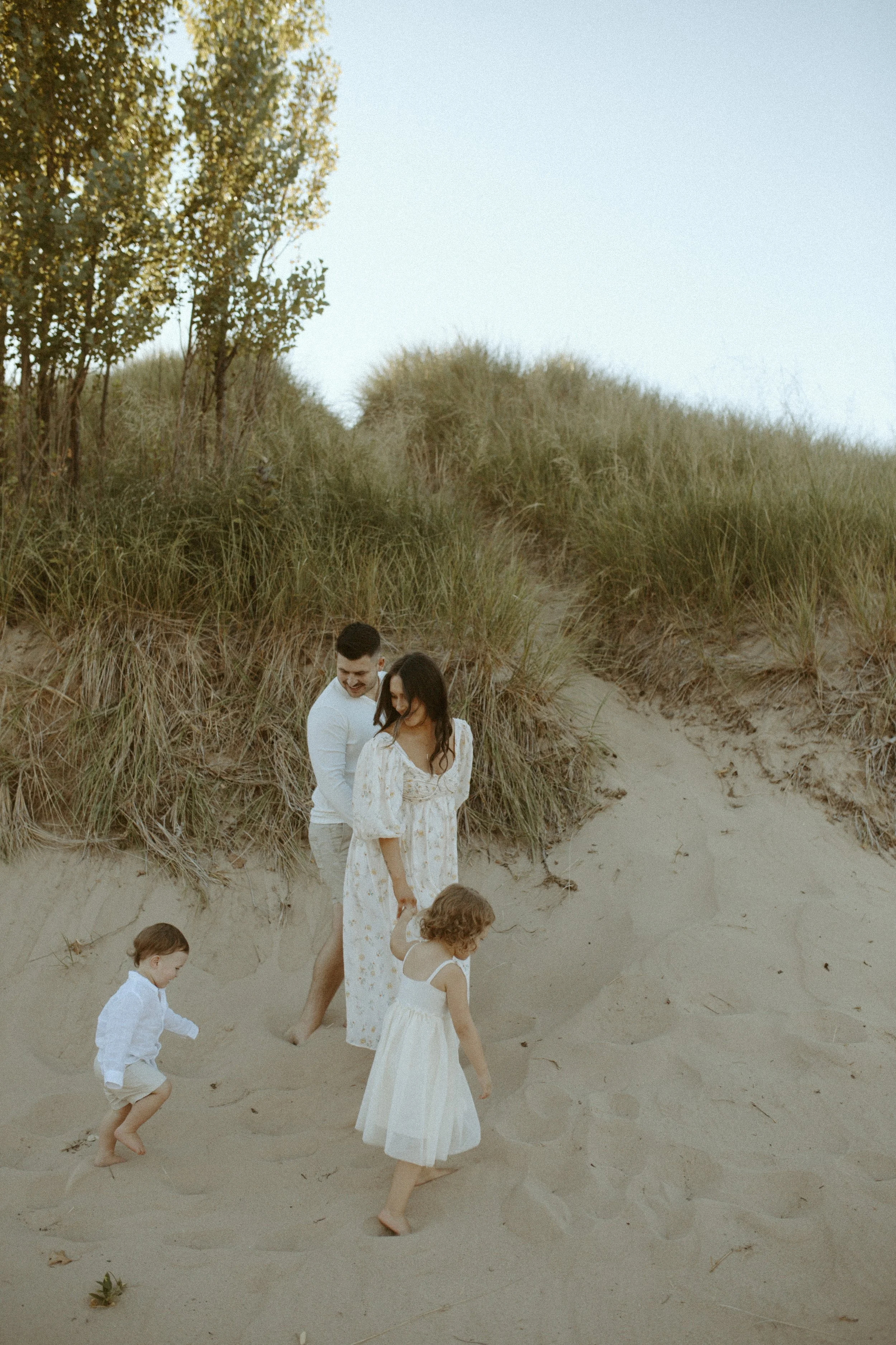 A family of five walking barefoot on a sandy beach, with dunes and grass in the background, during daytime.
