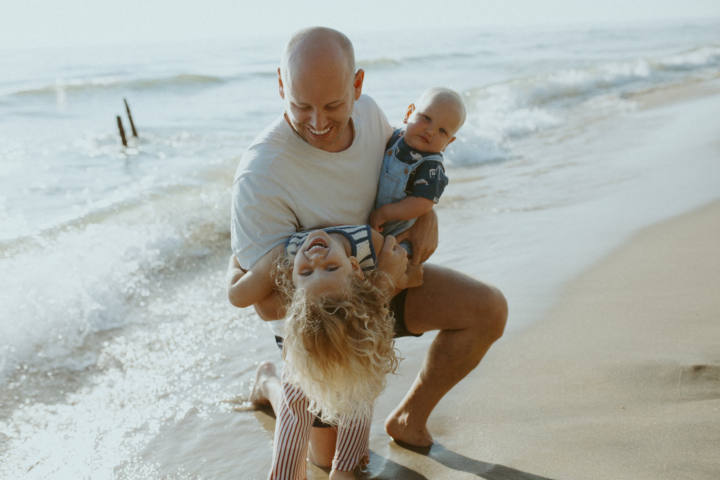 A man playing at the beach with two young children, one girl and one boy, near the shoreline with gentle waves in the background.