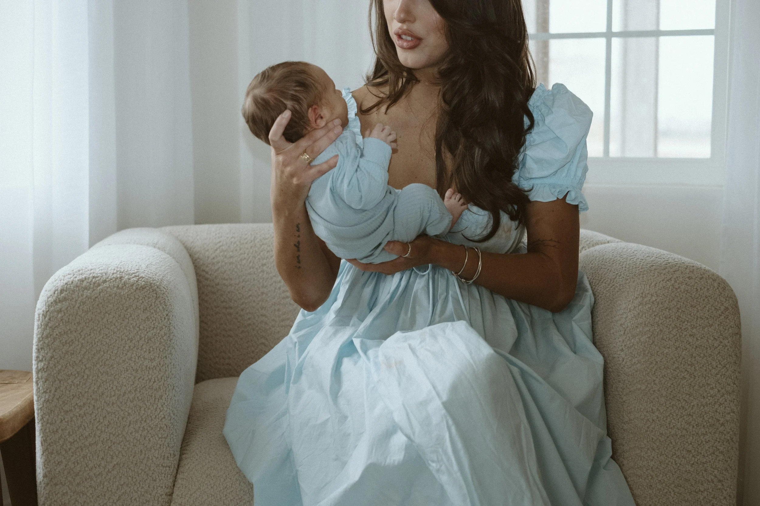 A woman holding a newborn baby in a bright room near a window with white curtains.
