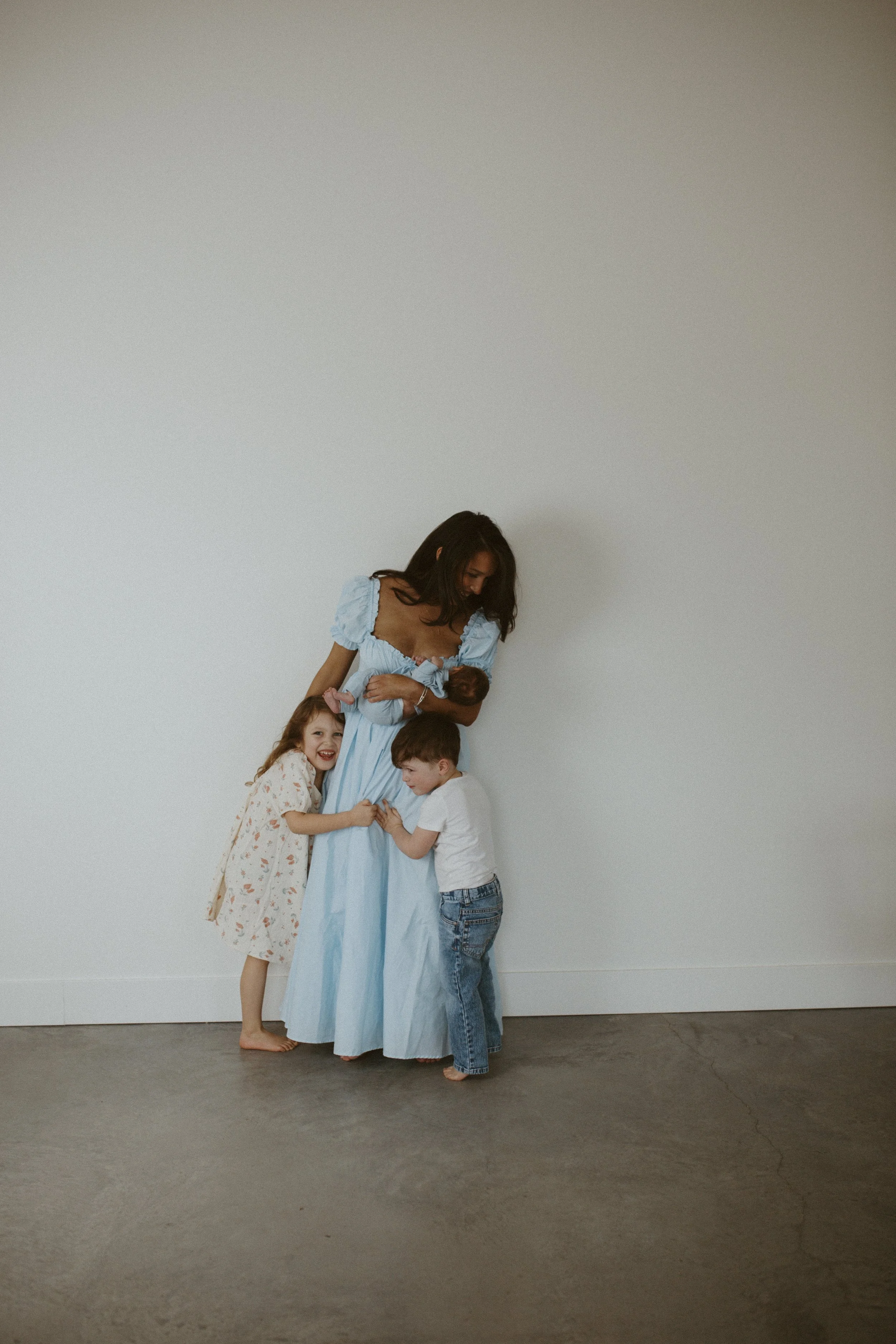 A woman in a light blue dress holding a newborn baby, with three children gathered around her, against a plain white wall.