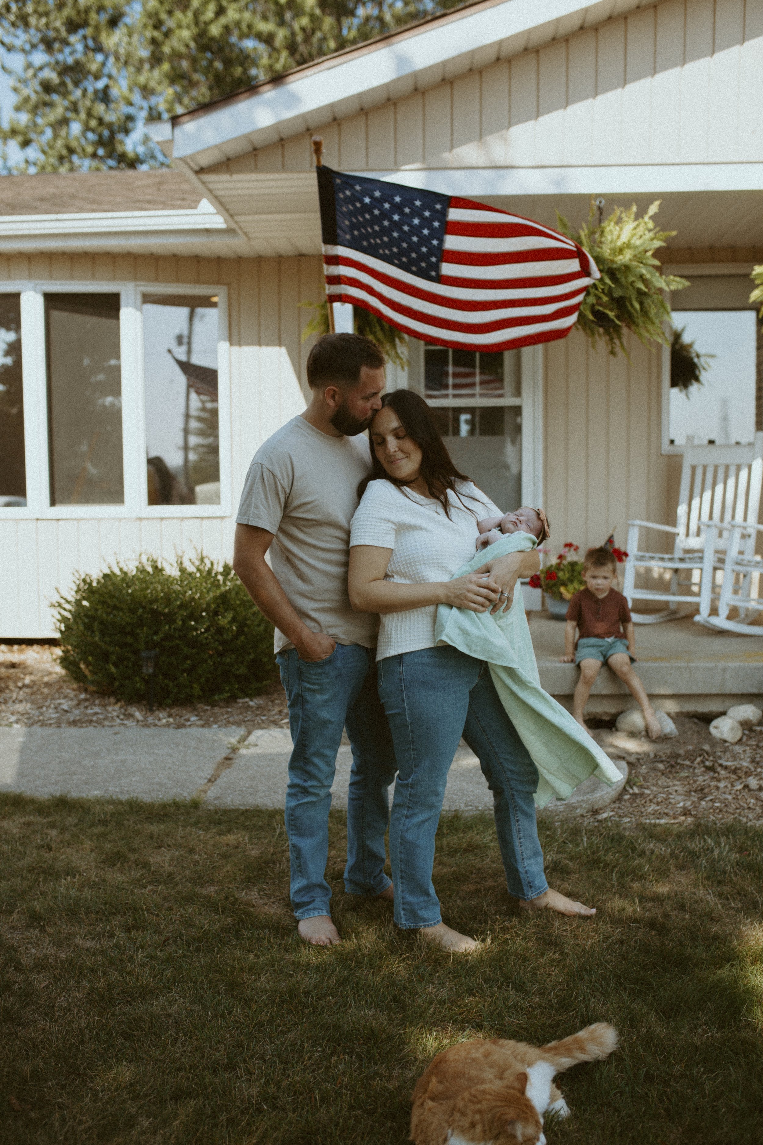 A couple with a newborn baby standing outside in front of a house decorated with an American flag and hanging plants, with children and a dog in the yard.