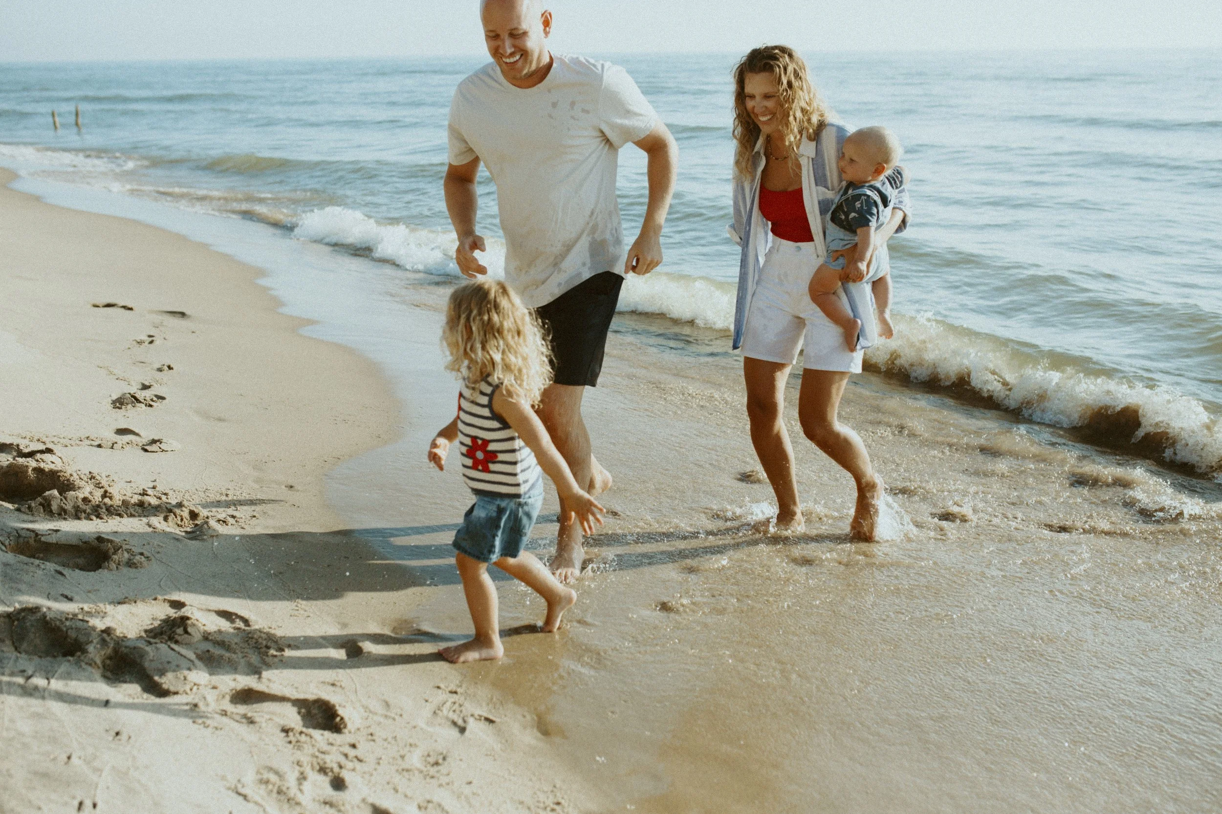 A family enjoying a day at the beach, with two adults and two young children, playing near the water's edge on a sandy beach.