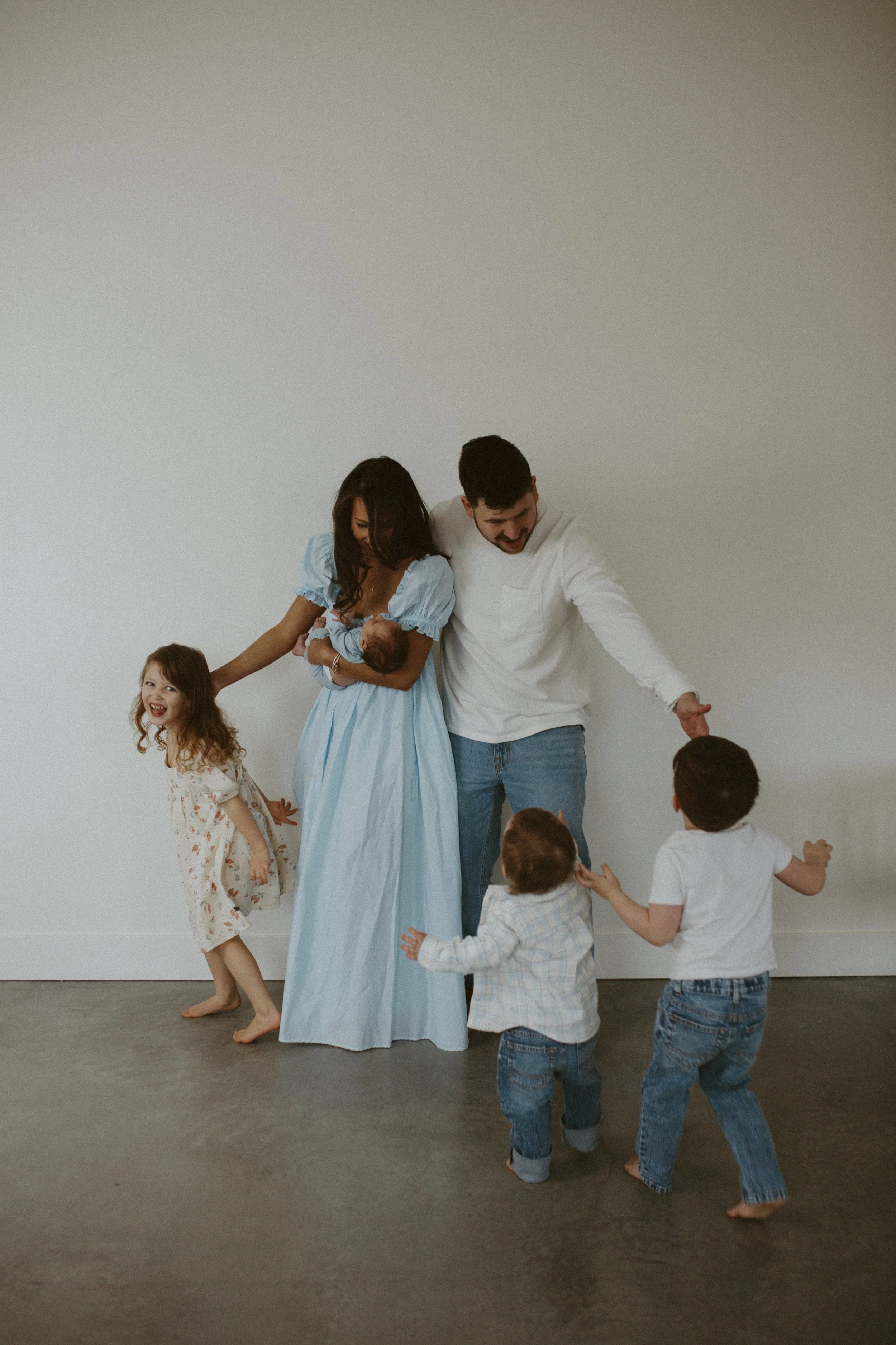 A family of five, including a woman, a man, and three children, is dancing and playing on a gray floor against a plain white wall. The woman is holding a baby, the man and children are interacting happily, with some barefoot and others in casual clot