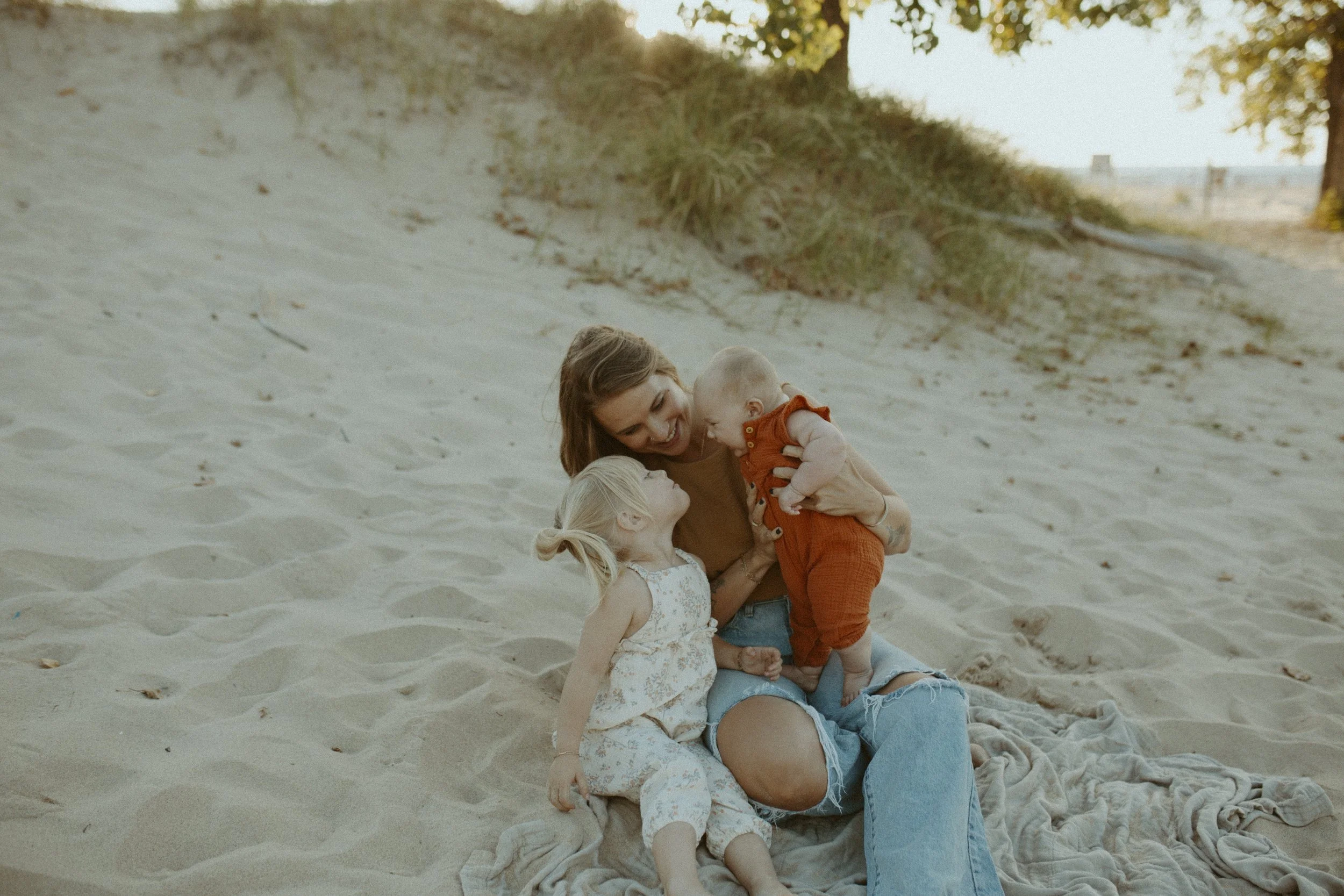 A woman with shoulder-length brown hair and a smile sits on the sand at a beach, holding a baby in an orange outfit. A young girl with blonde hair in a ponytail, wearing a white dress with floral patterns, sits nearby, holding the woman's hand. The s