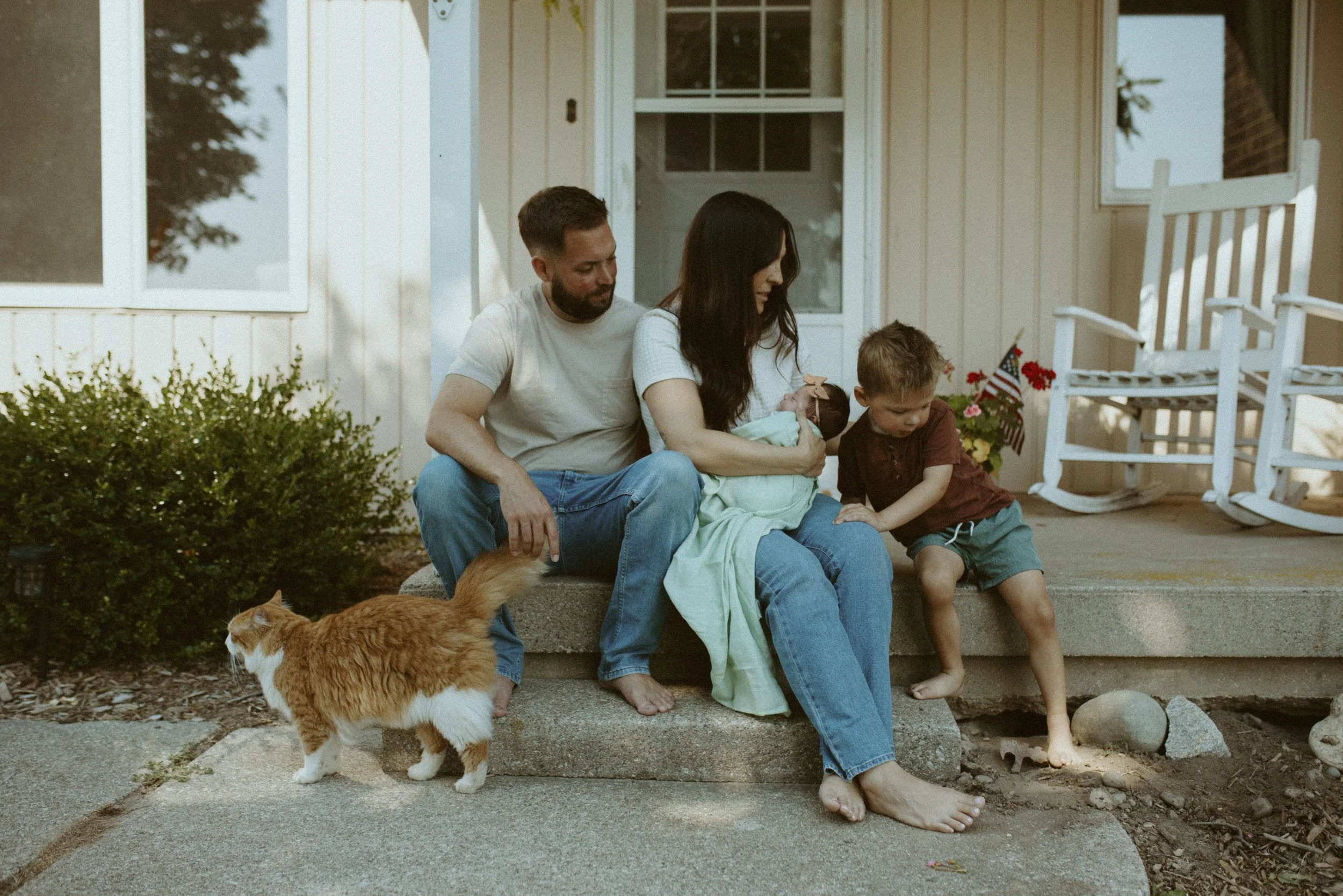 A family of four, including a man, woman, and two children, sitting on the steps outside a house with a white porch and a rock garden. An orange and white cat is walking nearby, and the family members are engaging in a moment of bonding.