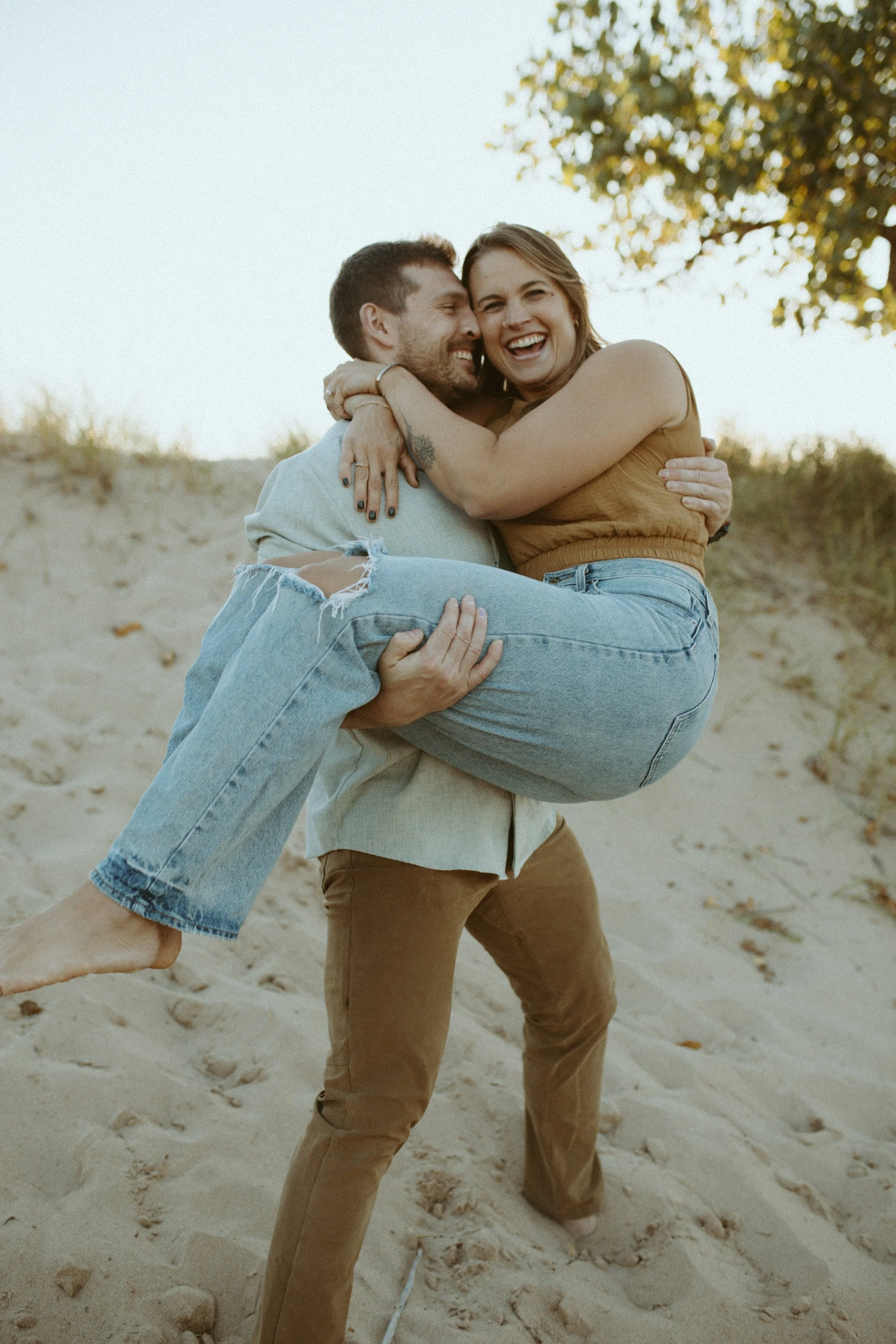 A couple on the beach, with the man lifting the woman in his arms. The woman has a big smile, and they are embracing each other happily. The background features sand and a tree with leaves.