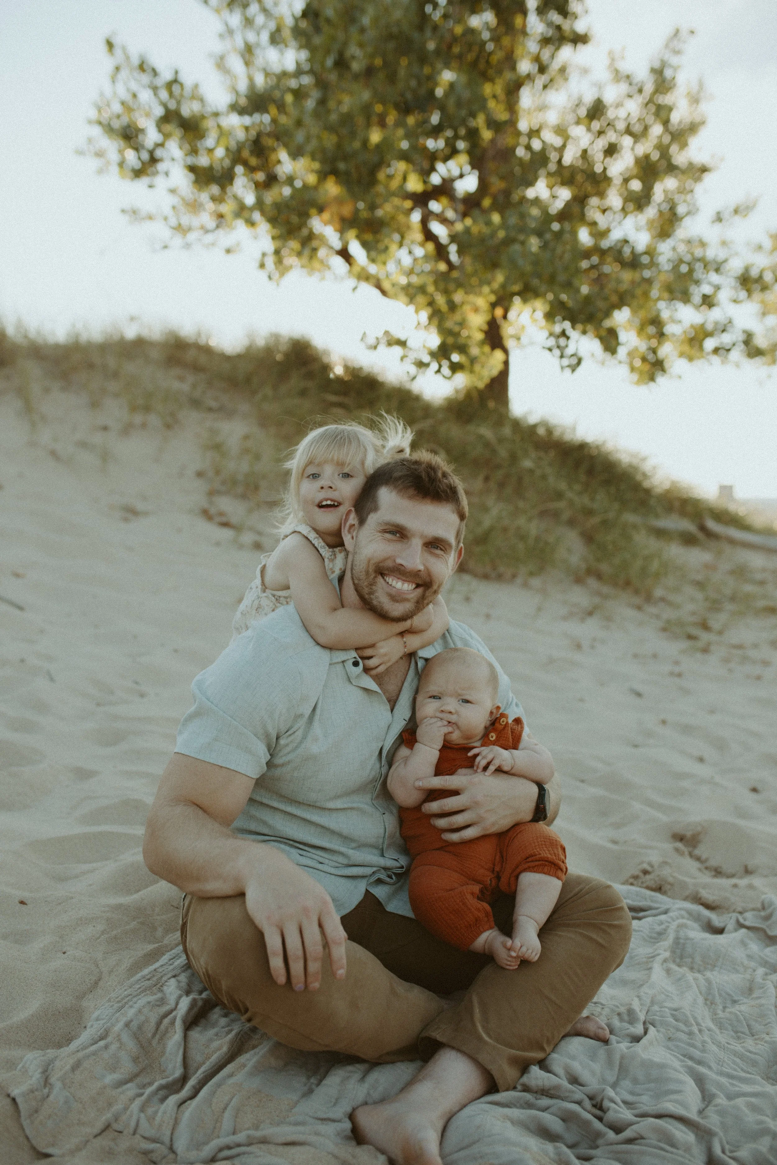 A man is sitting on a blanket on the sand in a beach setting, holding two small children, a girl on his shoulders and a boy in his lap. The girl has blonde hair and is smiling, while the boy has short hair, is wearing orange overalls, and appears to 