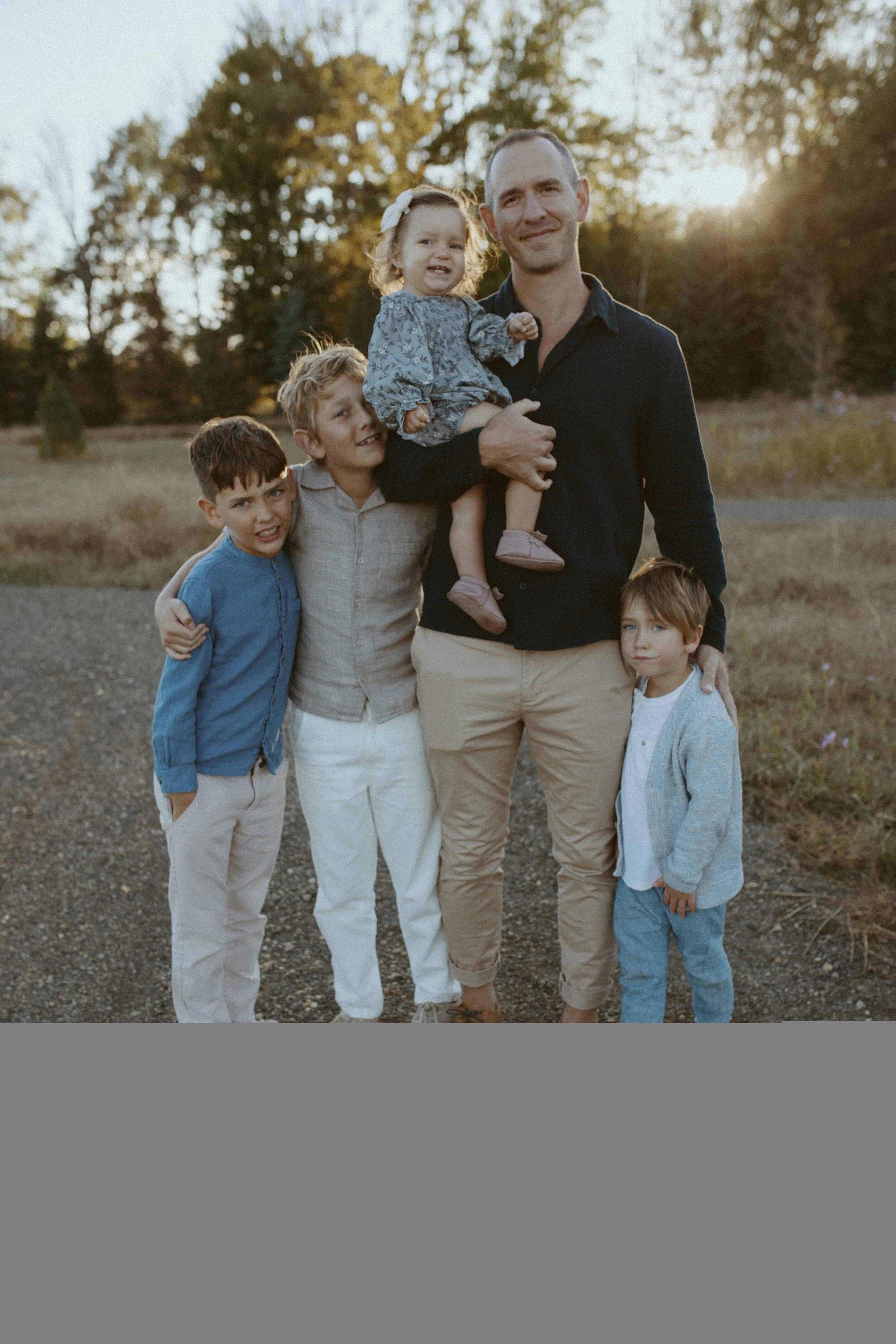 A man and five children outdoors at sunset, smiling and posing for a photo. The man is holding a young girl, and the children are standing on a gravel path with trees in the background.