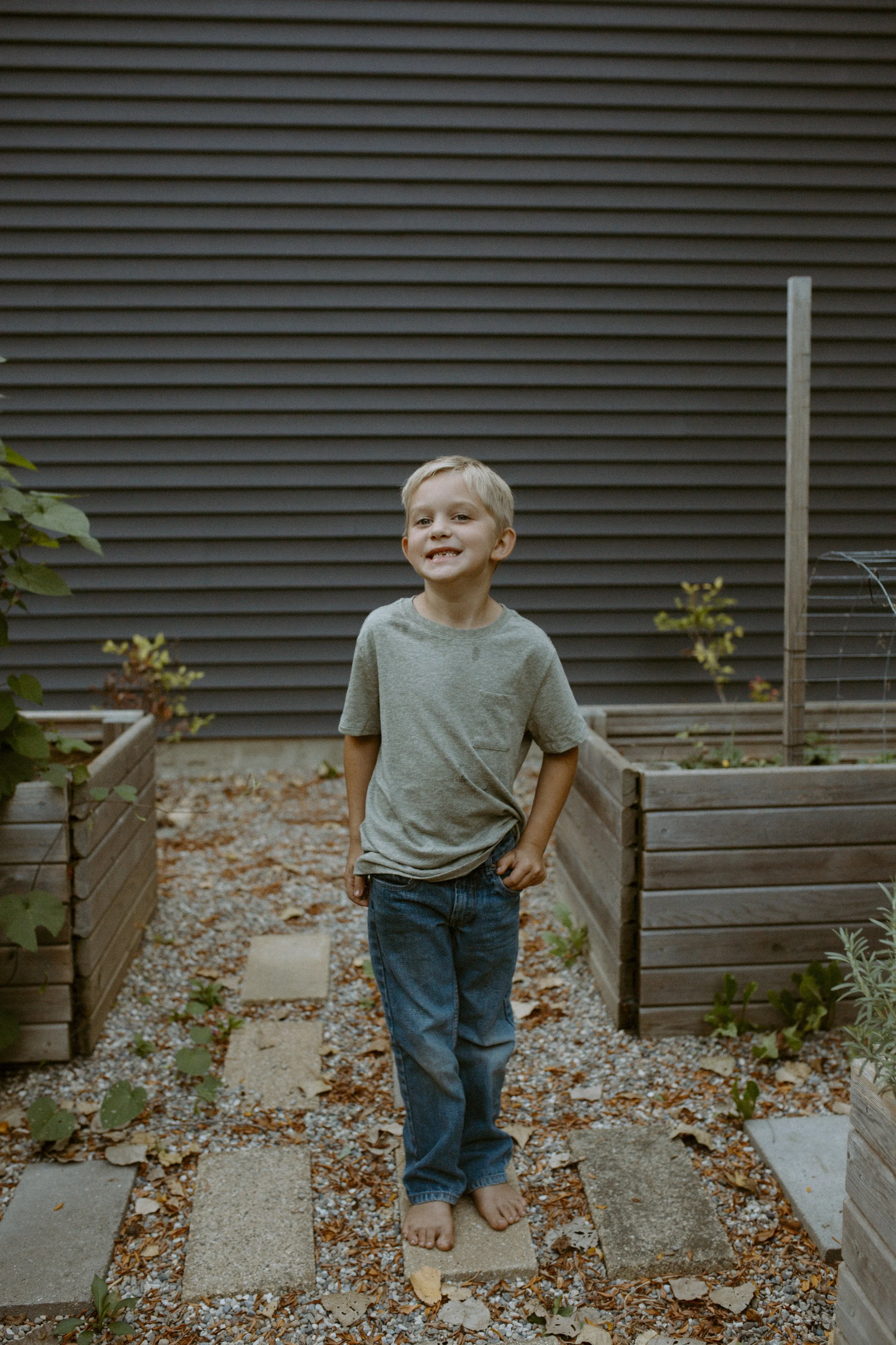 A young boy with blonde hair, wearing a gray t-shirt and blue jeans, standing barefoot on a stone path in a garden with wooden raised beds filled with plants, and a dark brown siding wall in the background.