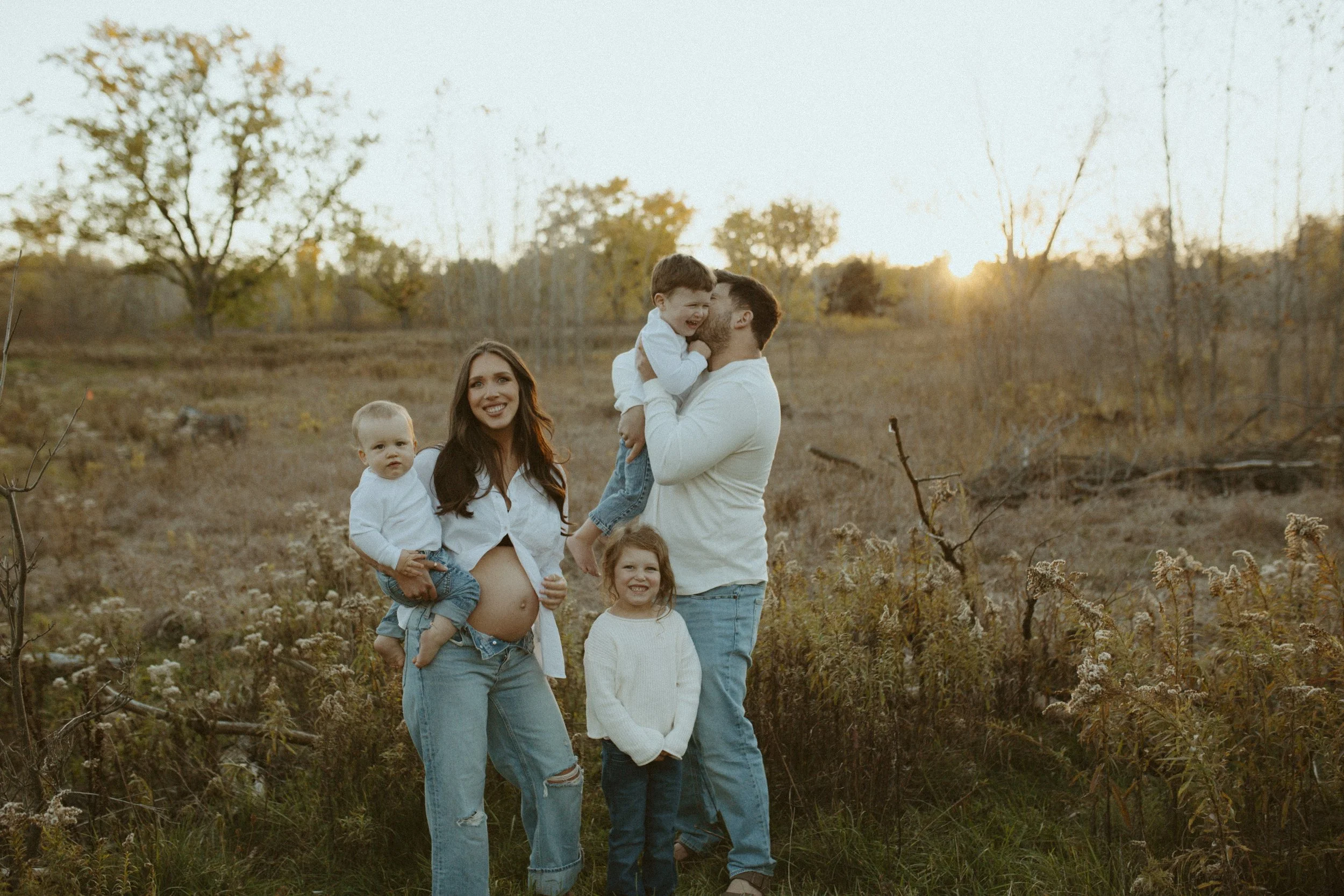 A family of five standing outdoors in a field during sunset, with trees in the background. The pregnant mother is holding a young girl, a man is holding a young boy, and two girls are standing on the ground. The family is smiling and enjoying the nat