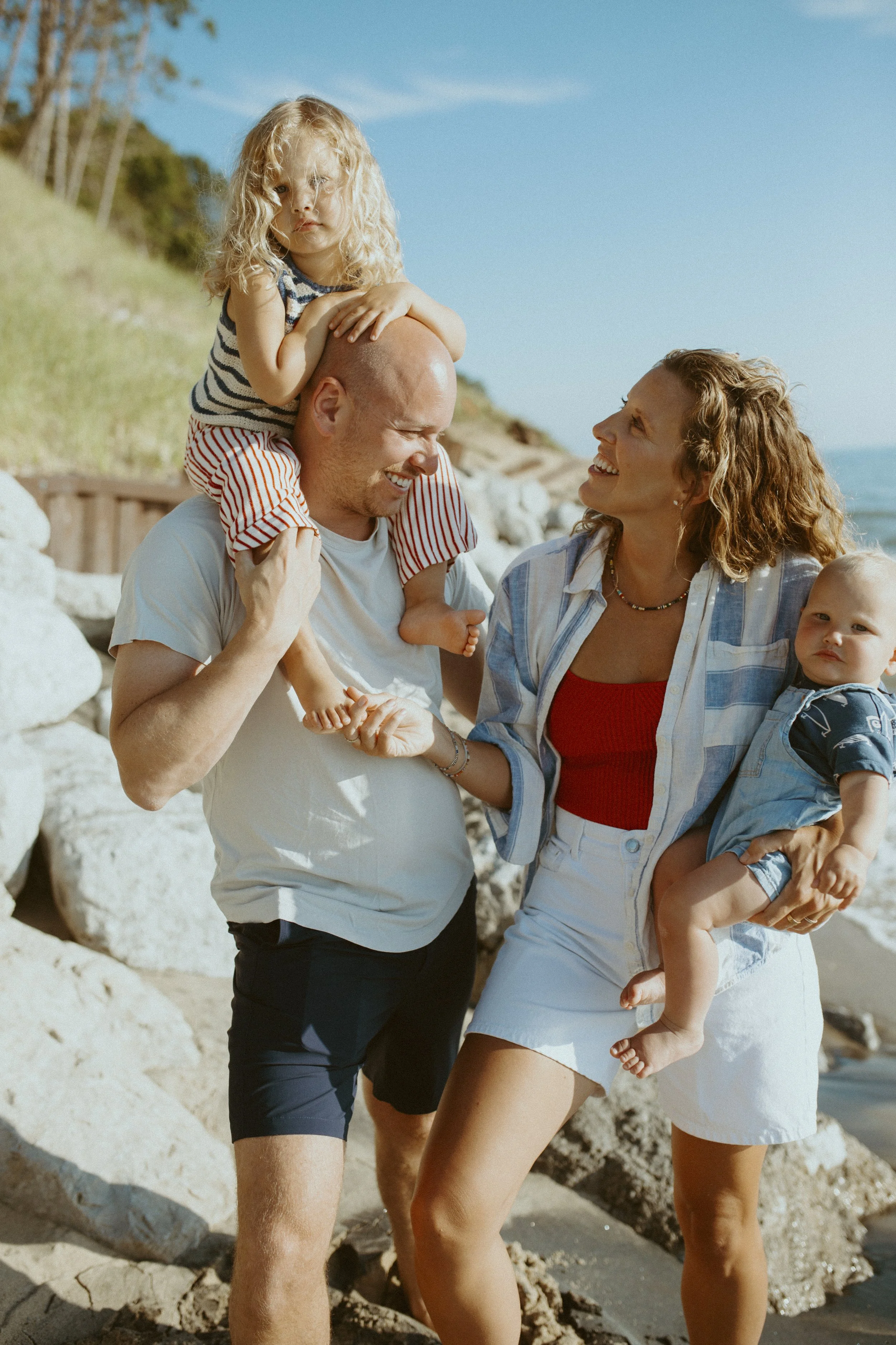 A family enjoying a day at the beach, with a young girl sitting on his father's shoulders, a woman smiling at the girl, and a toddler in her arms, all smiling and having fun.