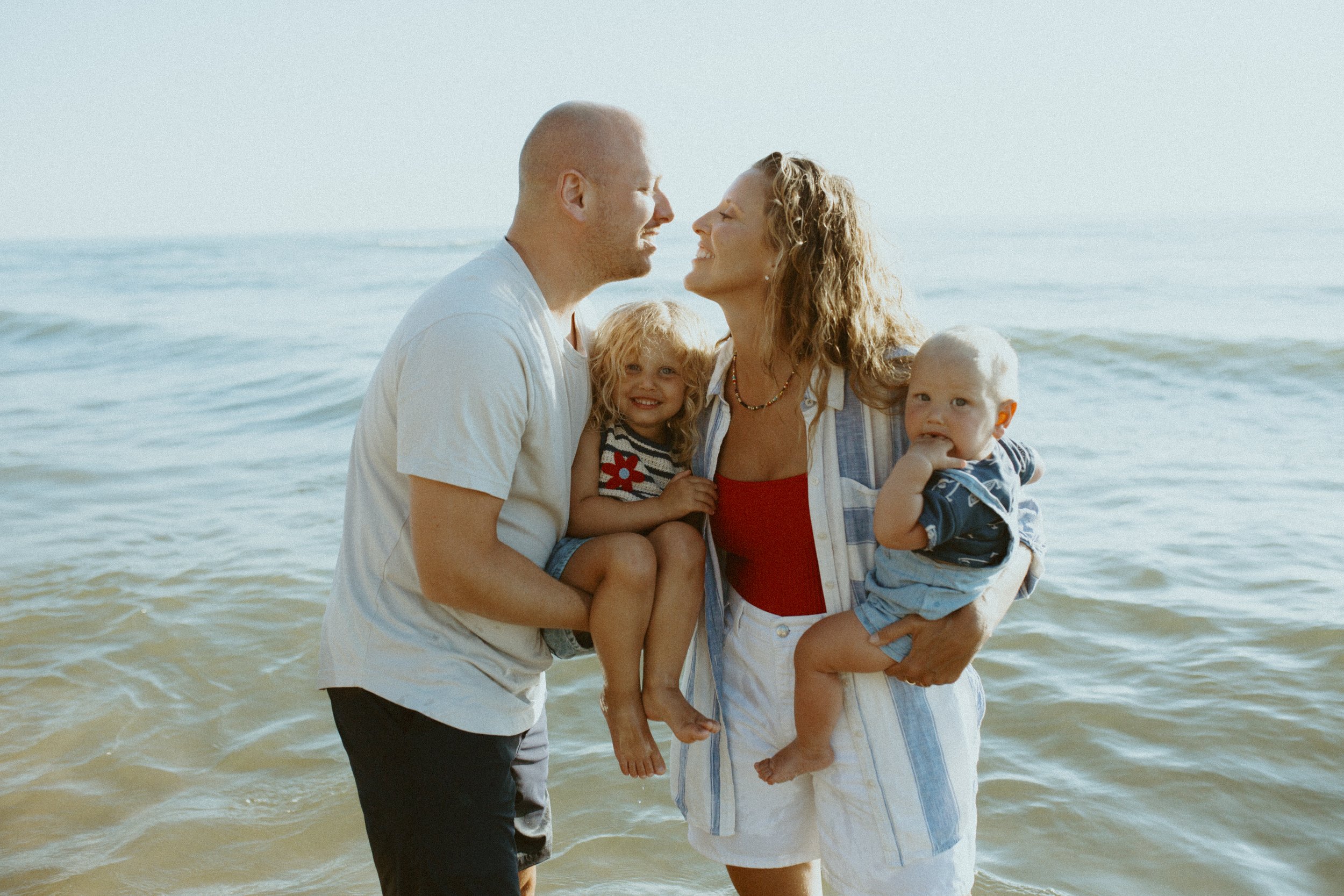 A family of four standing in ocean water, smiling and facing each other, with a clear sky behind them.