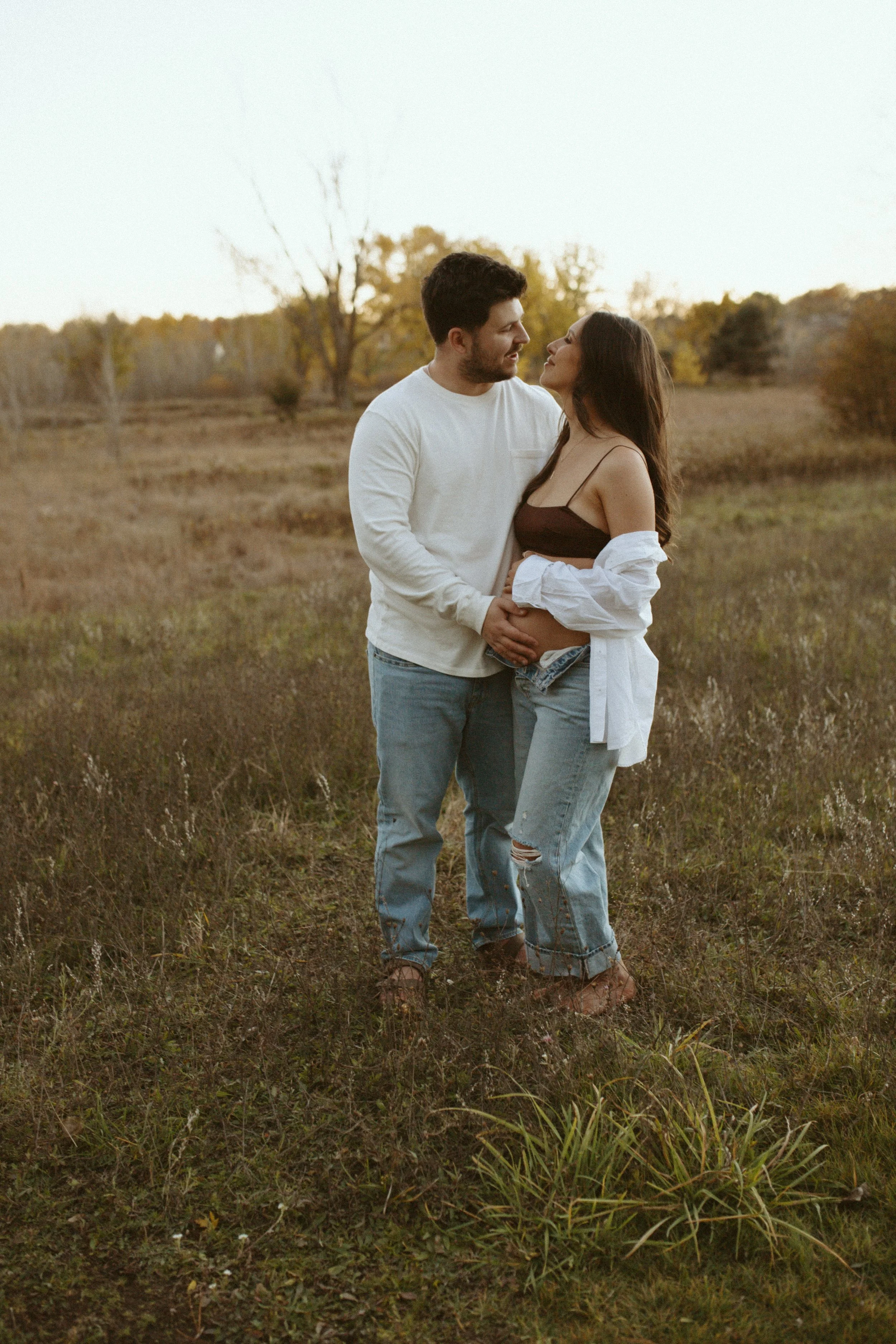 A couple standing closely in a grassy field during sunset, looking each other affectionately, with trees and open sky in the background.
