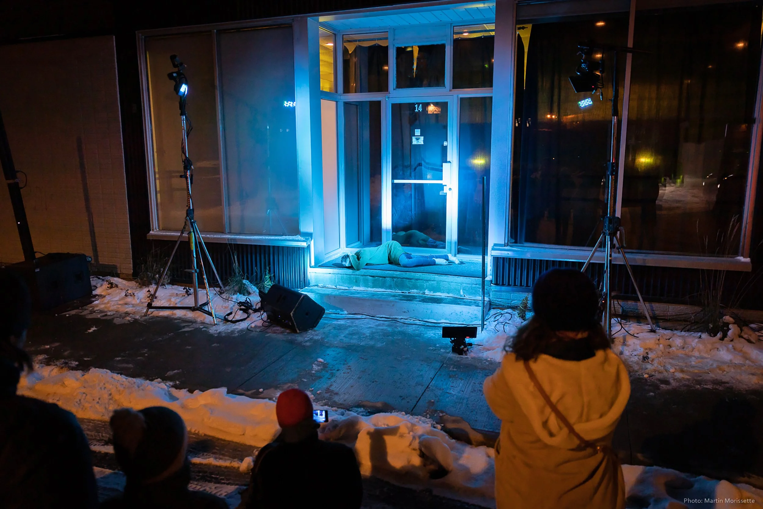 Nighttime scene of a woman lying face down on a small step outside a building, illuminated by blue stage lights. Several onlookers are watching, with some taking photos or videos. Snow is on the ground.