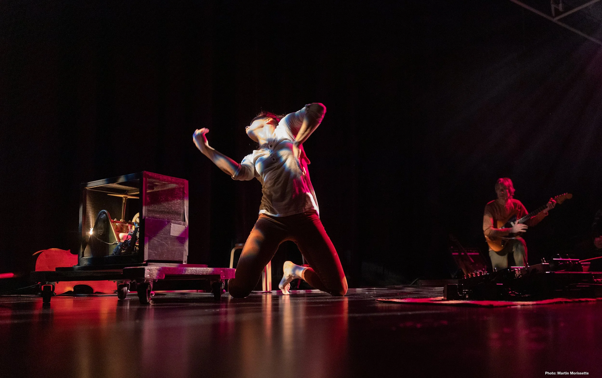 A performer is on stage with arms raised, surrounded by electronic equipment and a guitarist playing in the background with a Leslie Speaker, during a concert with dark lighting and red stage lights.