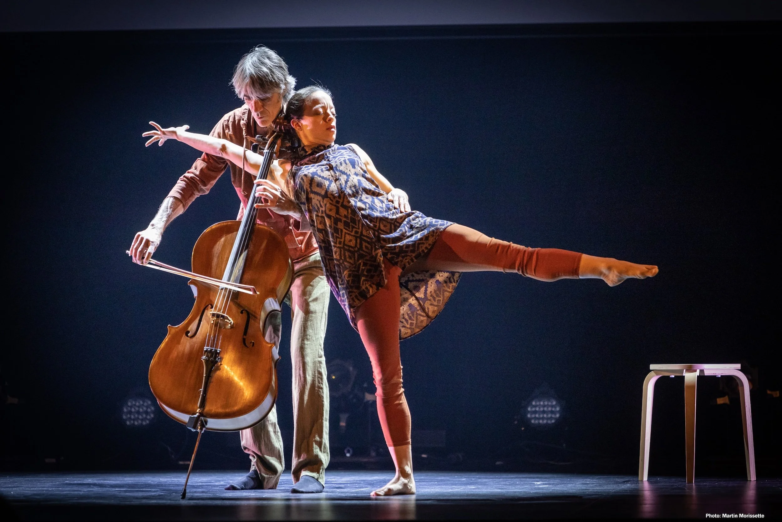 Two performers on stage, one playing a cello and the other balancing with one leg extended sideways, during a dance or theatrical performance.