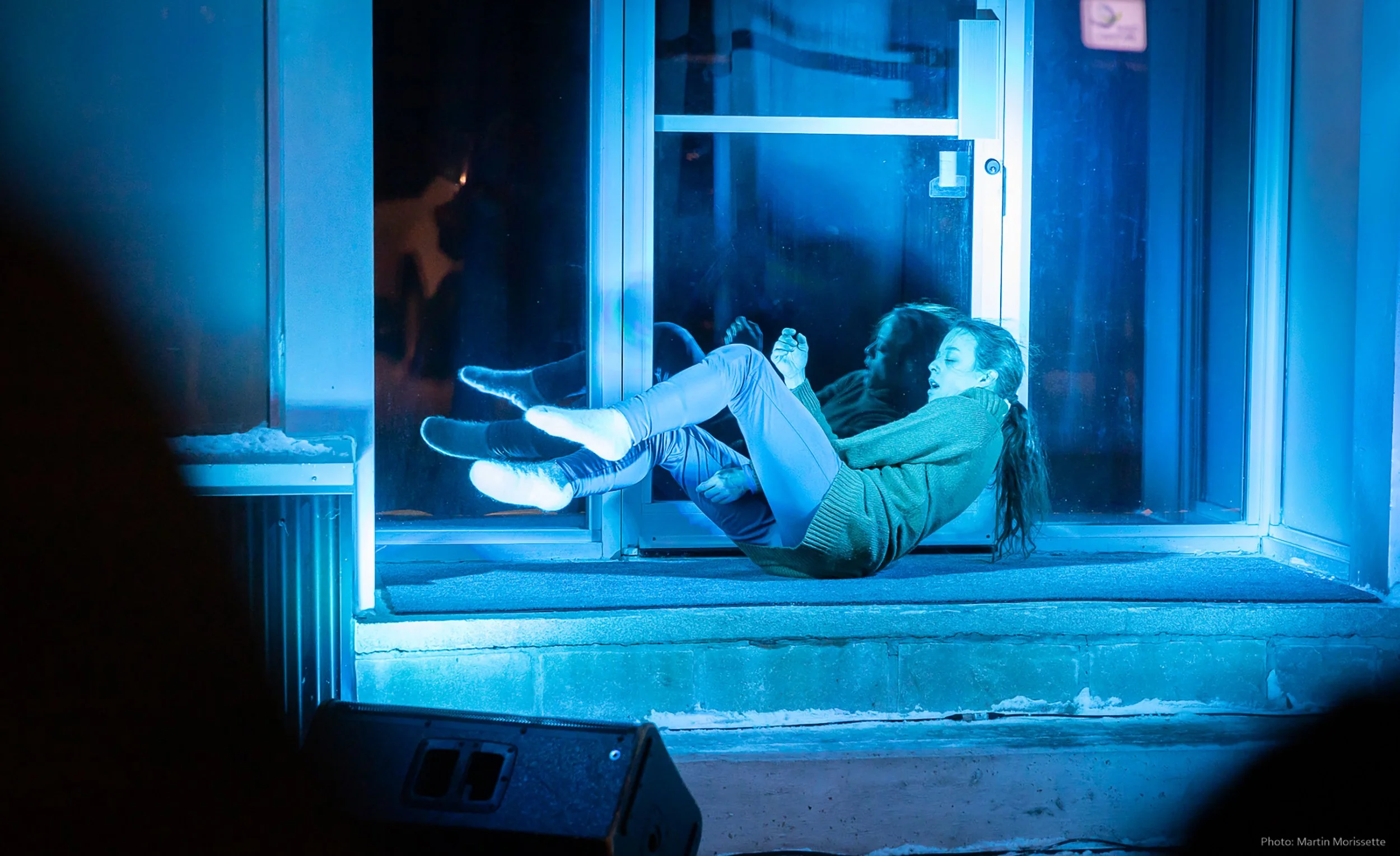 A woman in warm clothing is lying down on a small porch lluminated by blue light. She is performing dance outside during a cold winter.