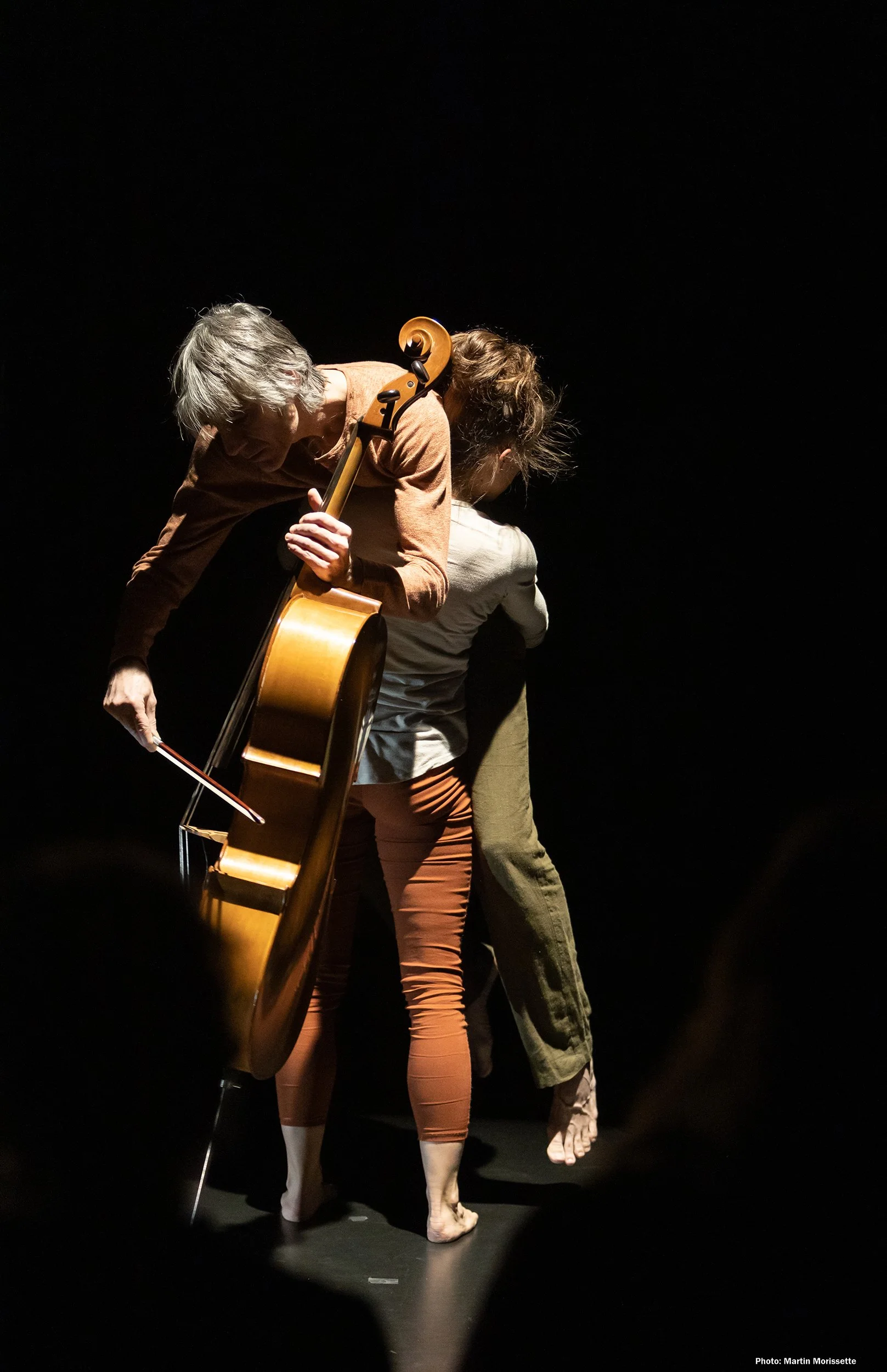 Two performers, one with a guitar and one without, are on stage dancing barefoot in front of a dark background.