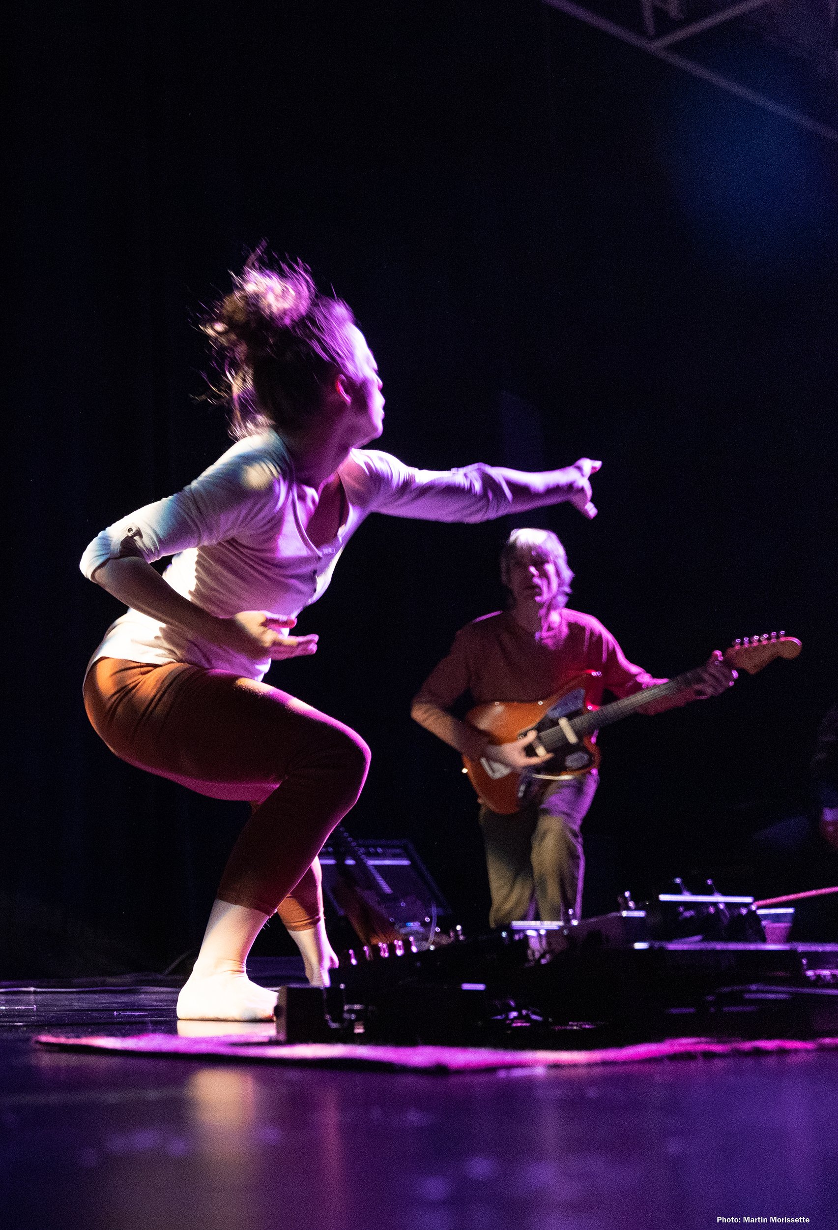 A woman with curly hair is dancing on stage with a guitarist playing in the background. The stage is lit with purple lighting.