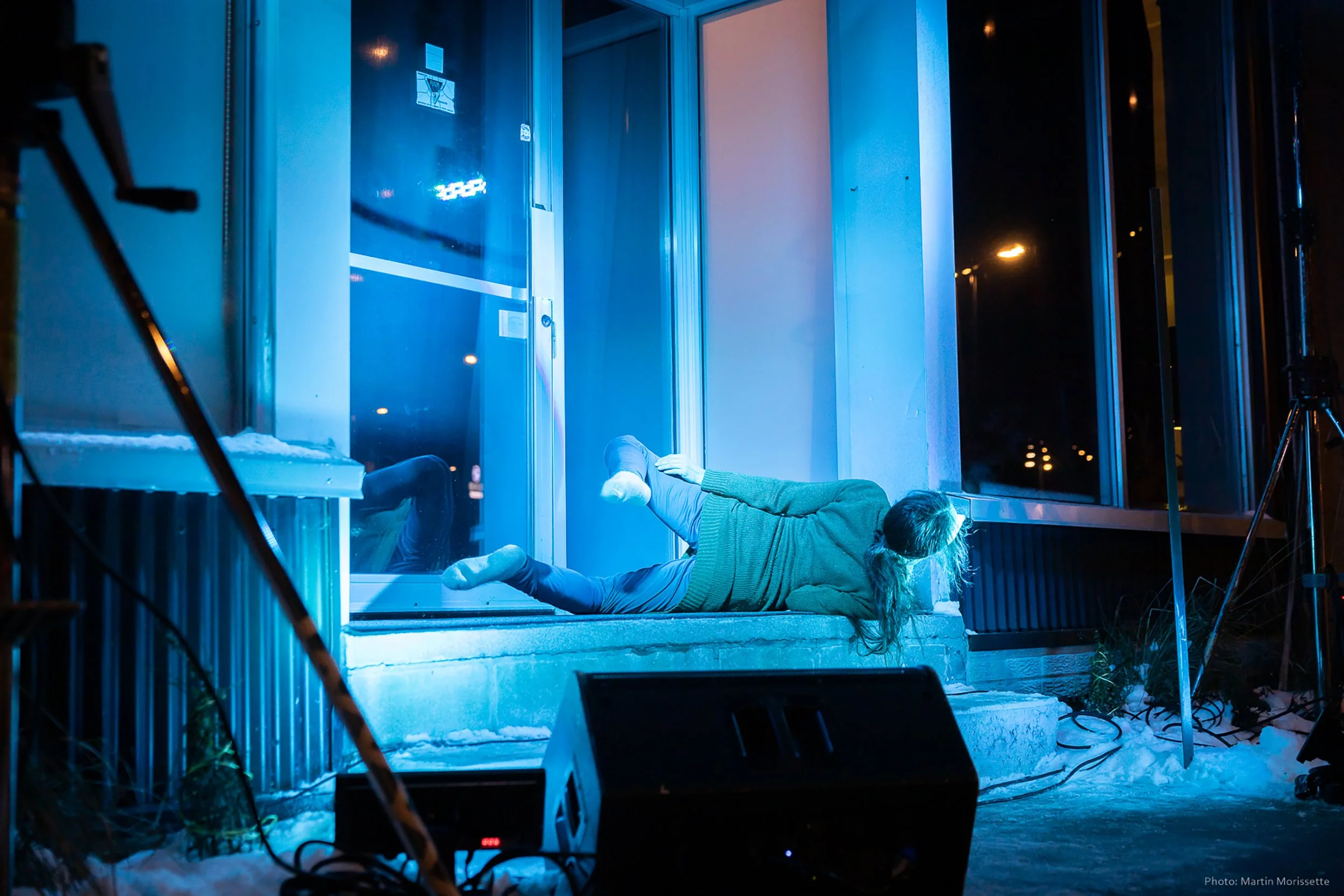 A young woman lying on the ground outside a building at night, illuminated by blue light, during a dance performance.