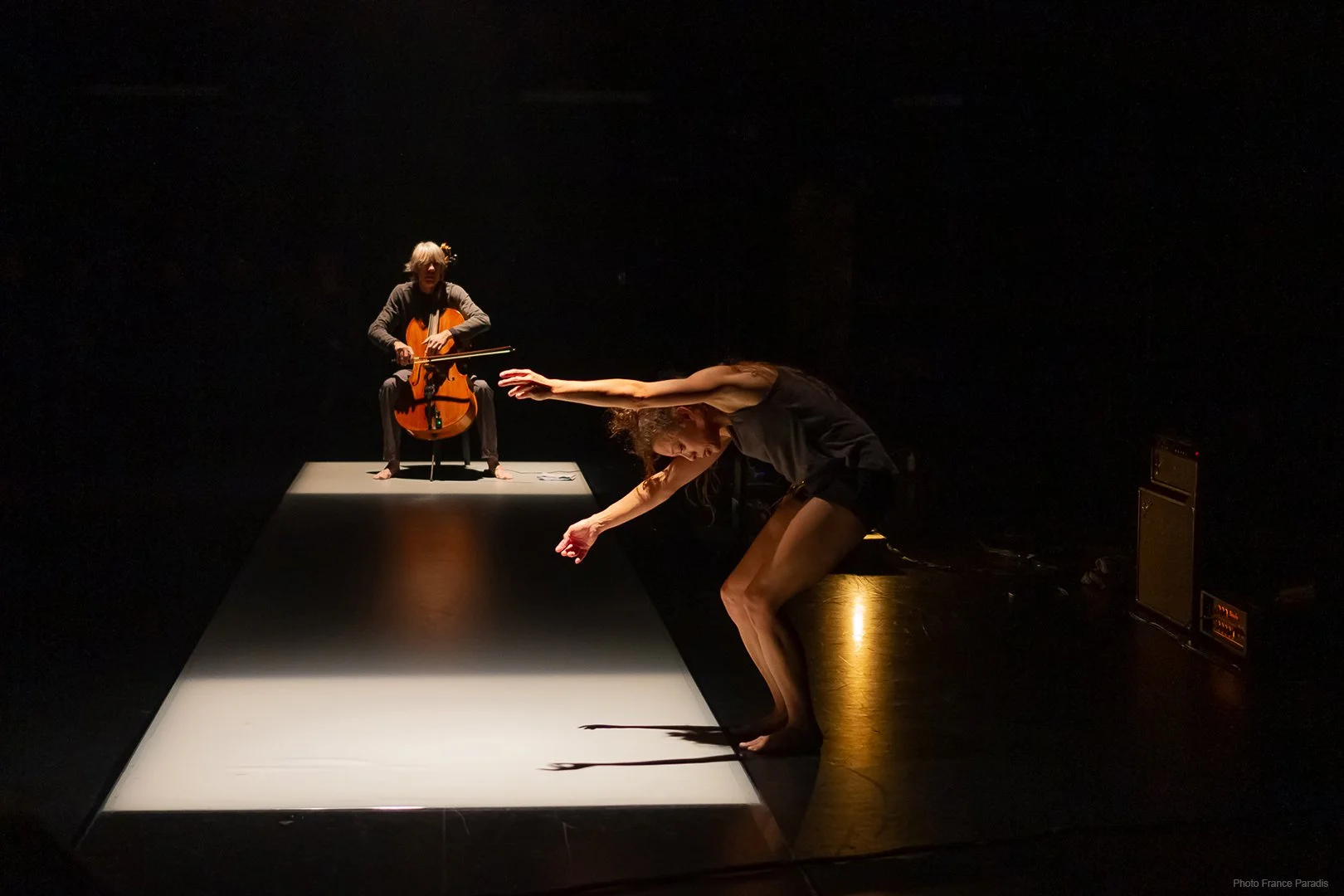 A performer crouching on stage with arms extended forward, and a cellist sitting on a platform in the background, playing the cello in a dimly lit theater.