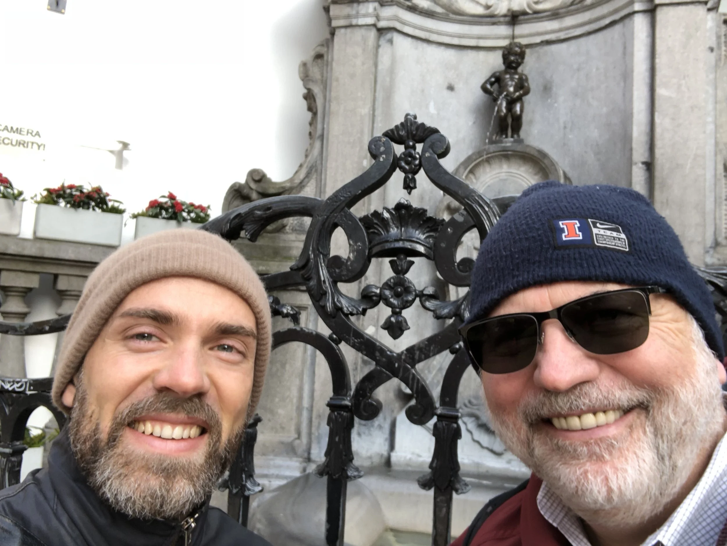 Twee mannen glimlachen voor een oude waterbron met beeldhouwwerken, een met een beige muts en de ander met een blauwe Nike hoed en zonnebril, in een historische stad.