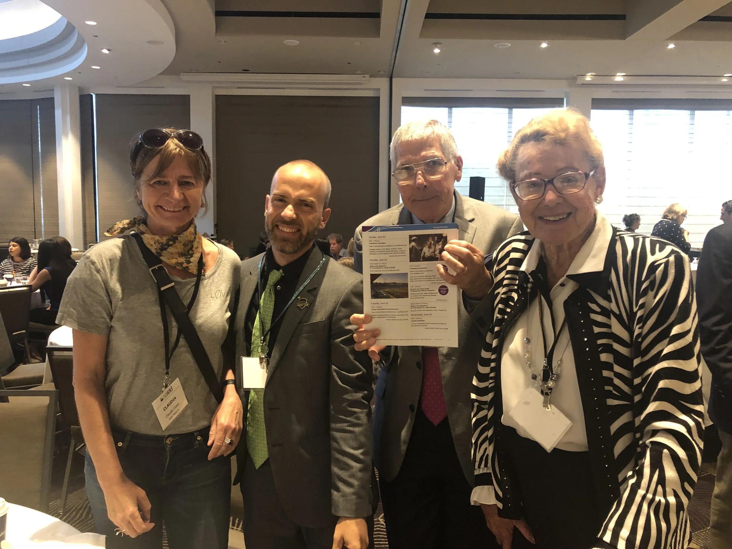 Vier mensen poseerden samen op een conferentie of bijeenkomst, twee vrouwen en twee mannen, in een zaal met andere aanwezigen op de achtergrond.