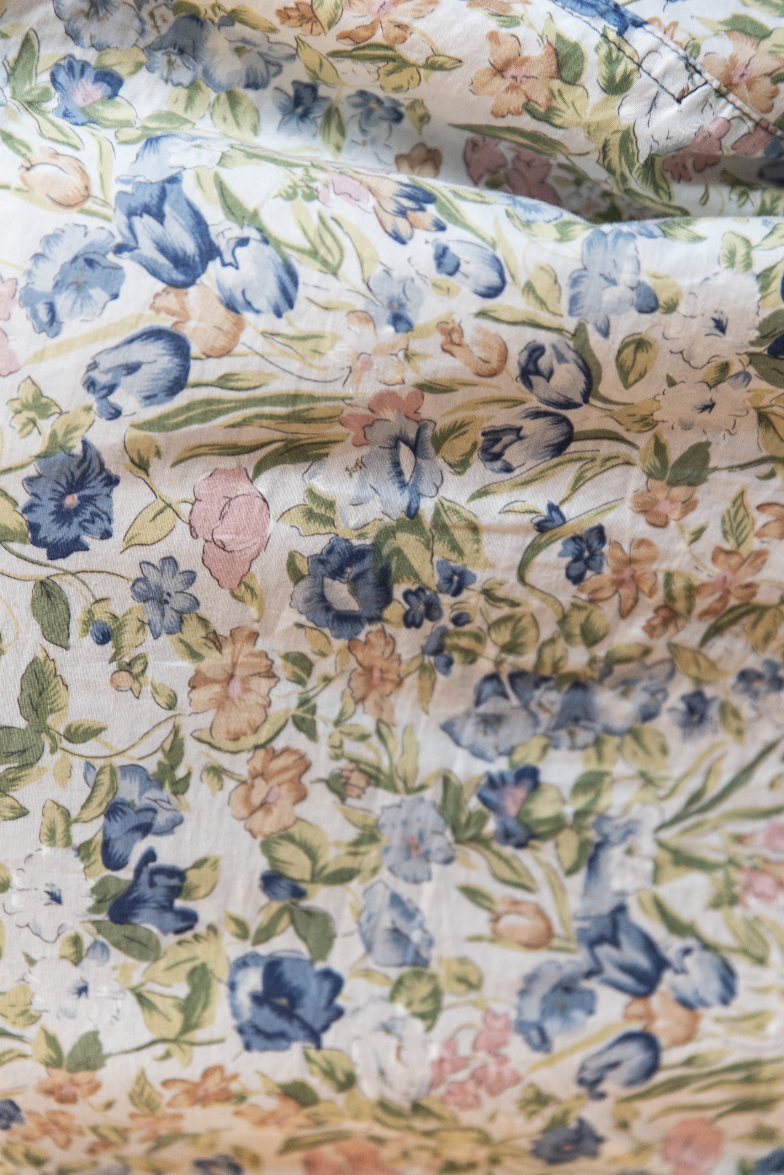 Close-up van een bloemrijk stoffen patroon met blauwe, roze en groene bloemen en bladeren op een lichte achtergrond.