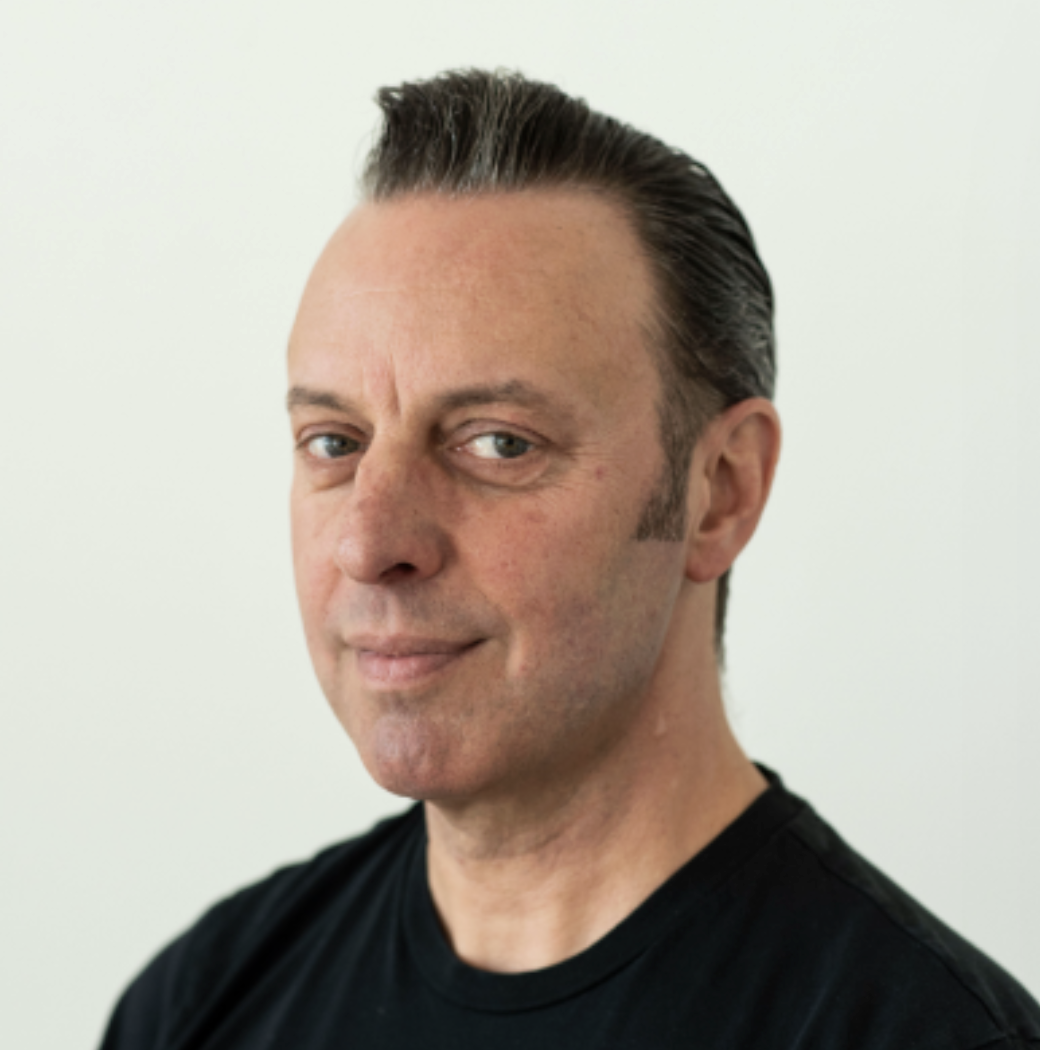 Portret van een man met donkere, steil gekamd haar en een lichte huid, lachend op een lichte achtergrond.