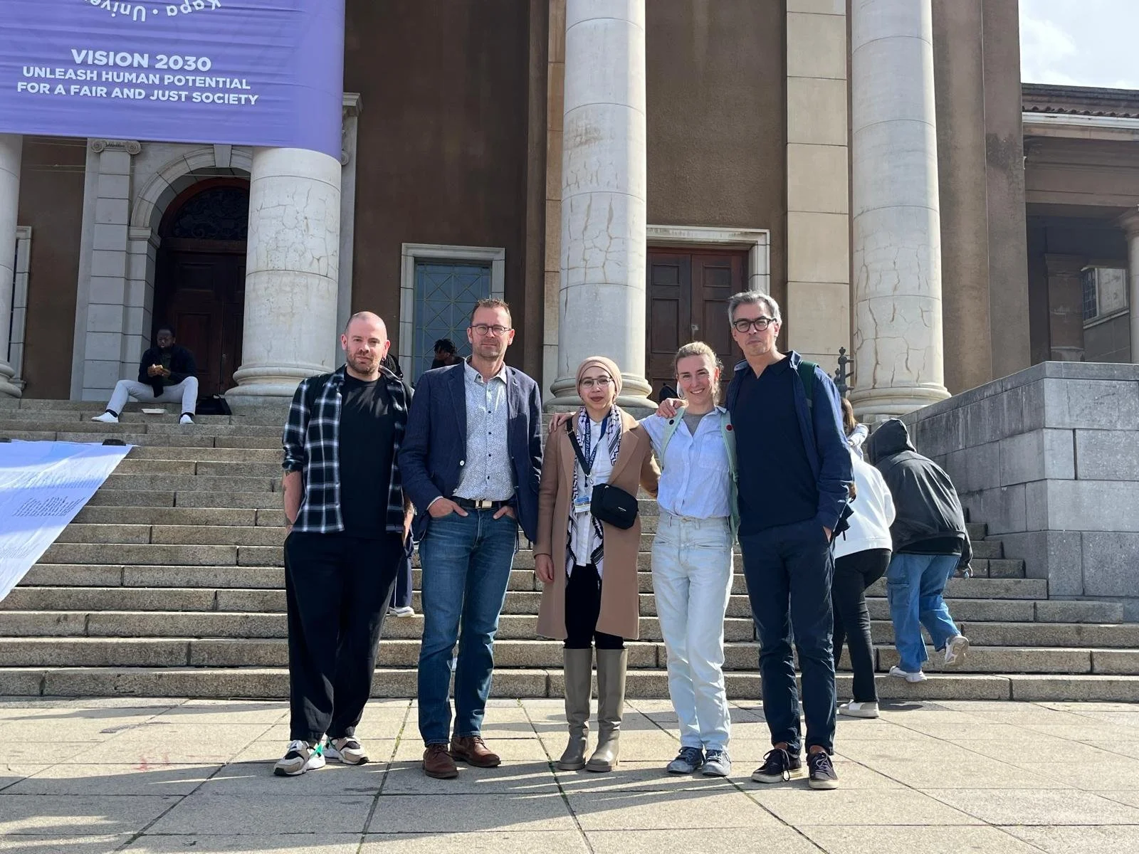 Vijf mensen staan samen voor de trappen van een grote, oude, stenen gebouw met grote zuilen, mogelijk een kerk of een belangrijke instelling. Ze poseren voor de foto en lachen, met enkele mensen op de achtergrond.