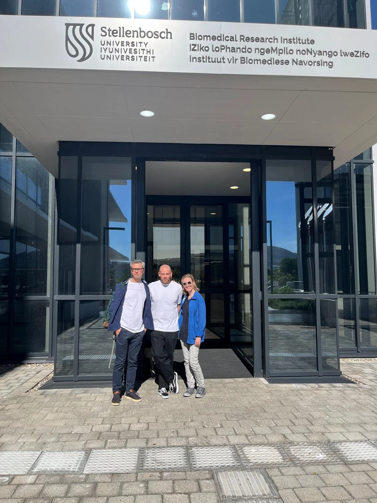 Drie mensen staan voor het ingangsgebouw van het Stellenbosch University, het Biomedical Research Institute, op een zonnige dag.