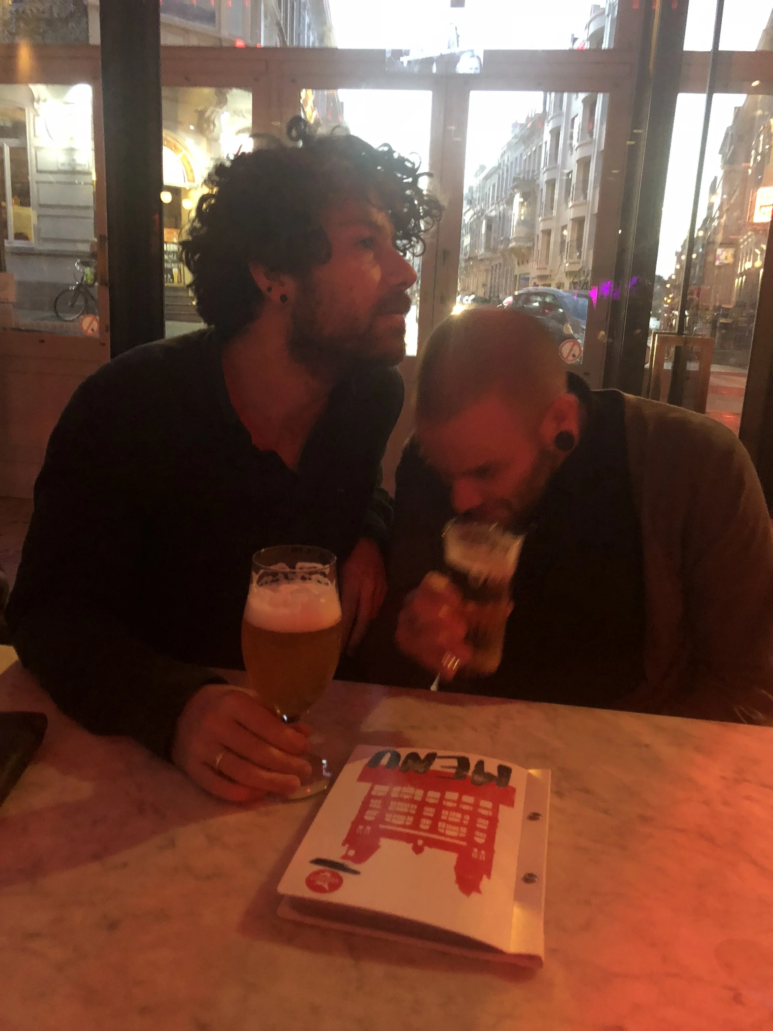 Twee mannen zitten binnen in een bar, ze hebben allebei een glas bier voor zich op de tafel. Eén man met krullend haar kijkt naar buiten terwijl de ander met een kaal hoofd drinkt, het menu ligt op tafel.