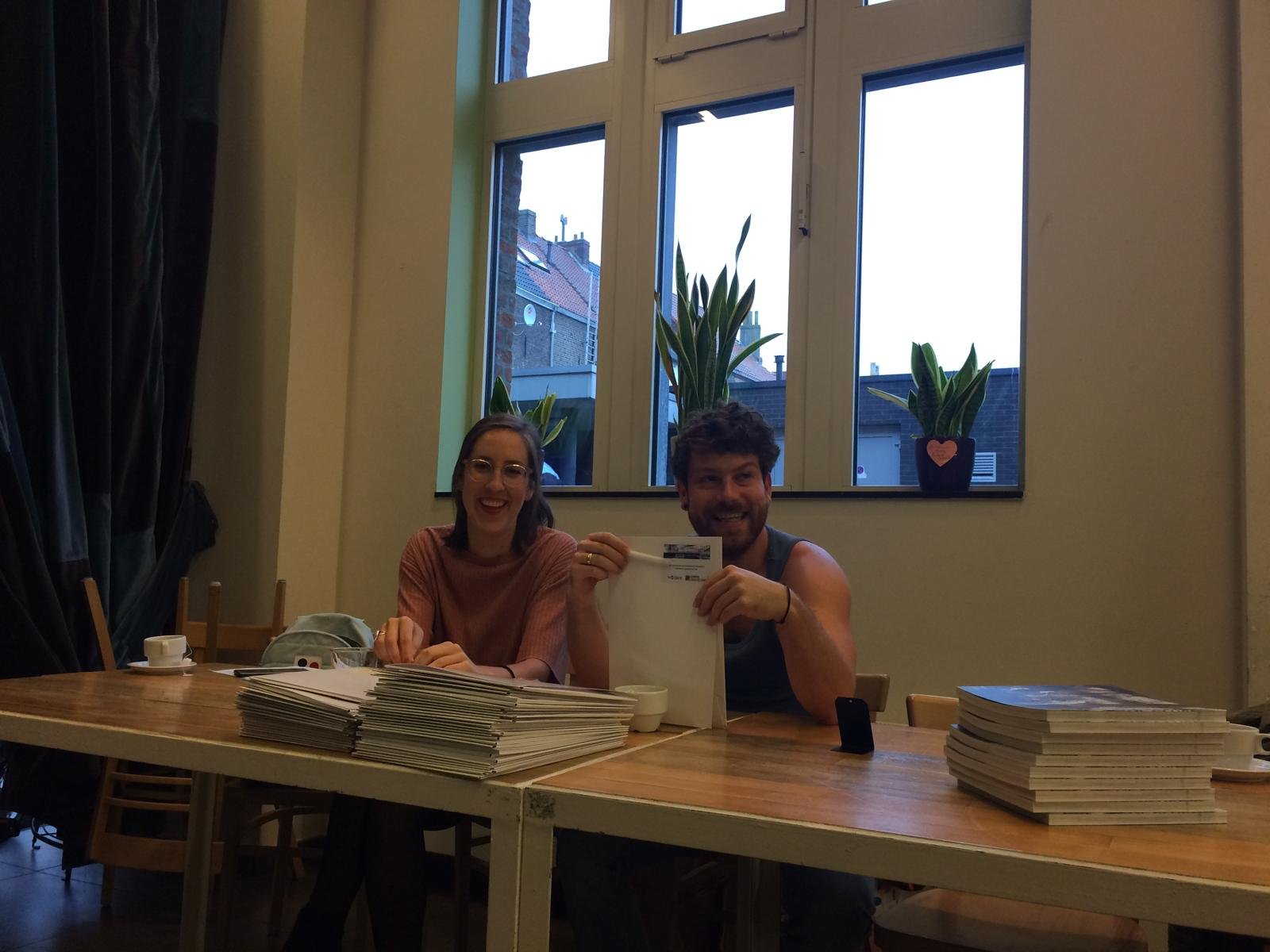 Twee mensen zitten aan een tafel in een kamer, lachen en tonen een brief of een document. Op de tafel liggen stapels documenten en kleine schaaltjes.