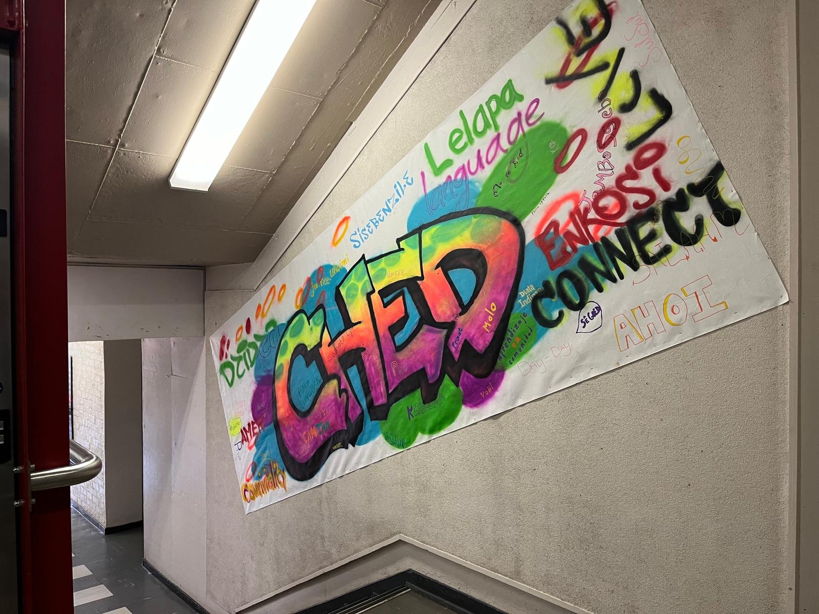 Een grote kleurrijke muurkunst met de woorden 'CONNECTED' en verschillende woorden in verschillende talen en kleuren eromheen, aan een betonnen muur in een binnenruimte.