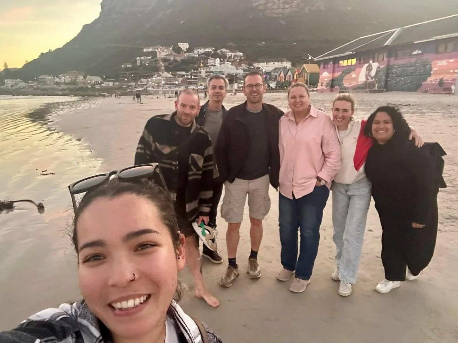 Een groep mensen poseren samen op het strand met een berg en huizen op de achtergrond. Het lijkt avond te zijn met een ondergaande zon.