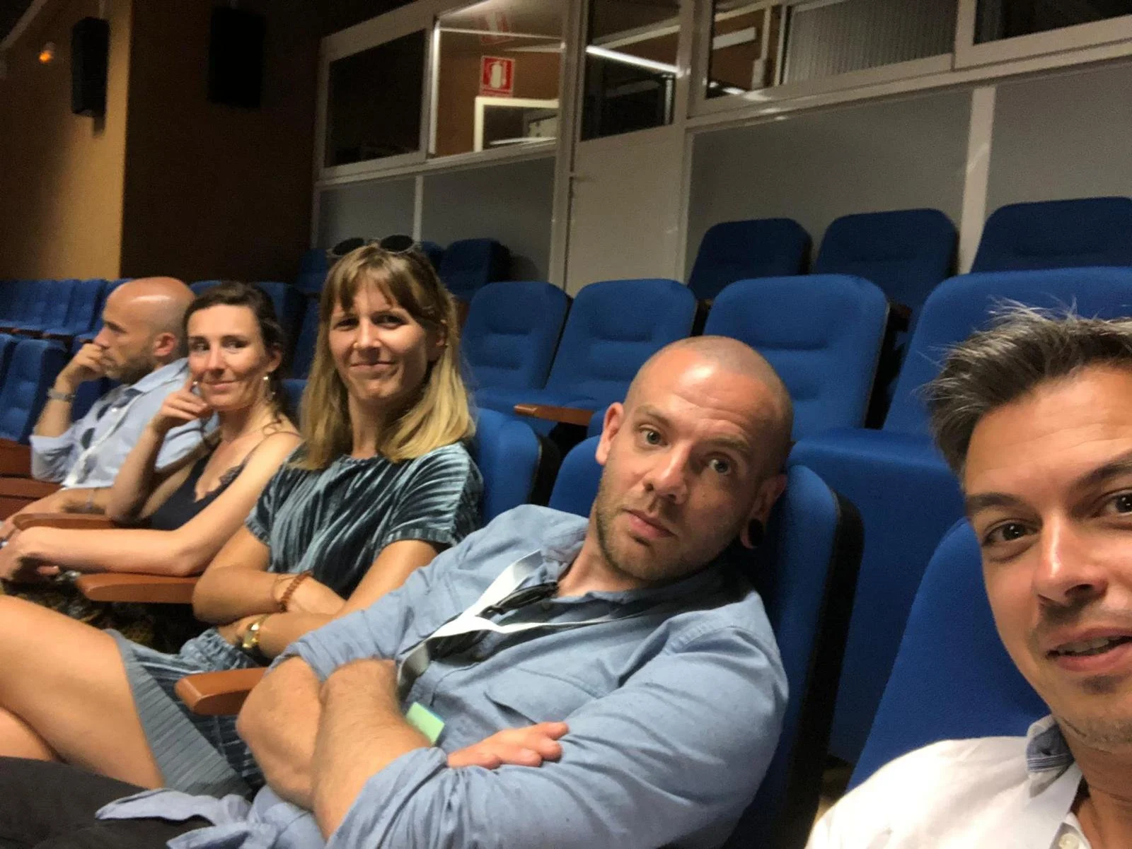Vijf mensen zitten in een zaal met blauwe stoelen, sommige kijken naar de camera, anderen niet.