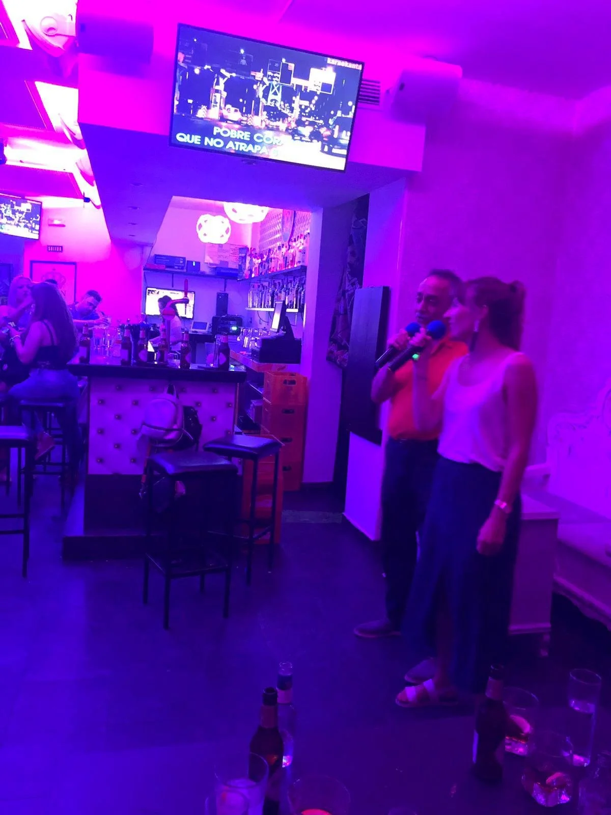 Binnen in een bar of pub met neonverlichting, mensen zitten aan de bar en aan tafels. Twee mensen met microfoons staan te zingen of te praten.