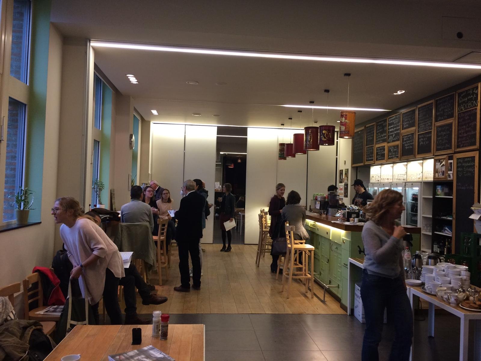 Drukke café met mensen die praten en werken, barista achter de counter, menu op de muren, tafels met kopjes en borden, grote ramen met planten.