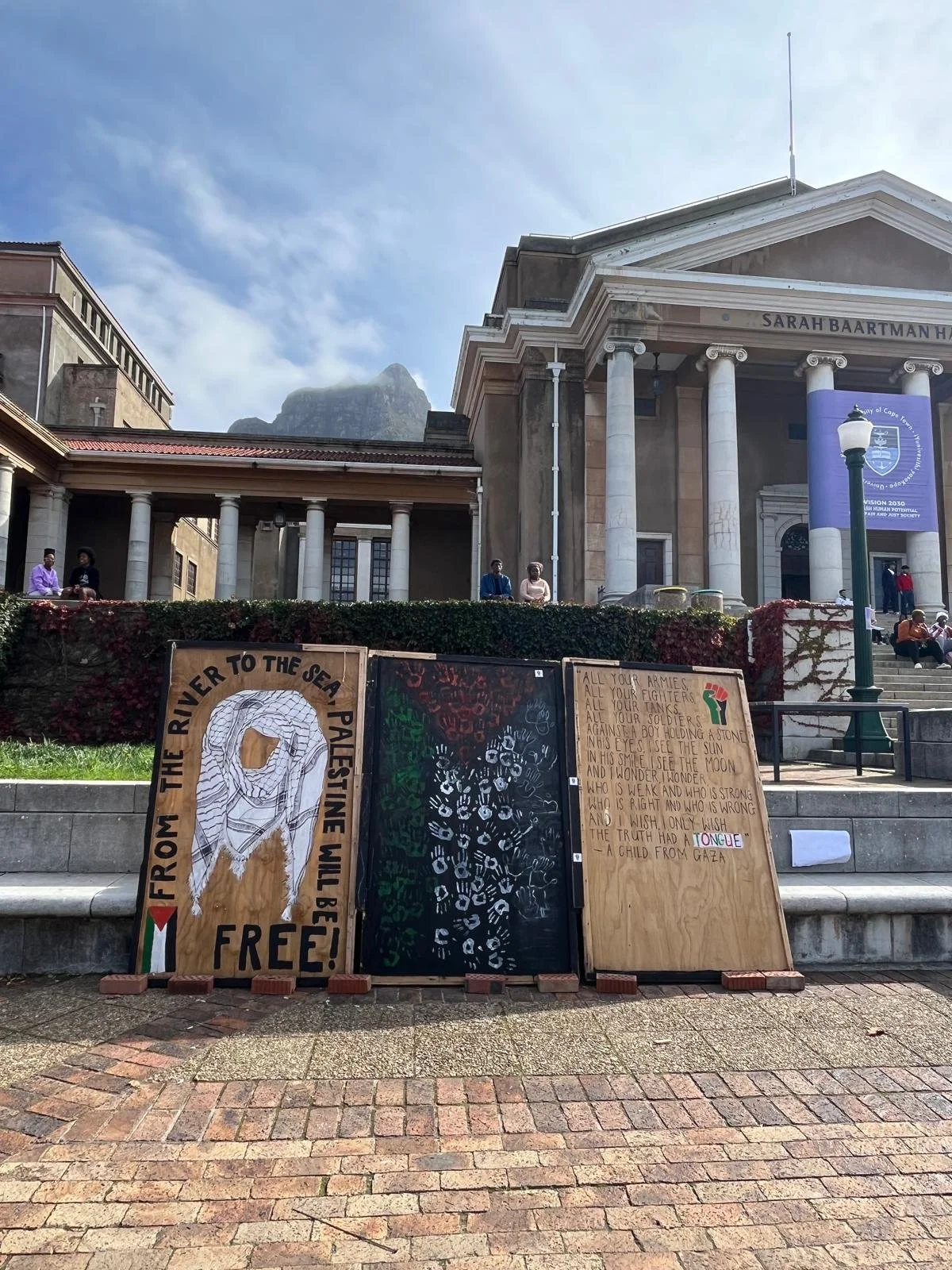 Drie posters staand op een trappenhuis voor het biertheater met een classical gebouw op de achtergrond en een berg. De posters bevatten teksten en tekeningen over Palestina en vrede.